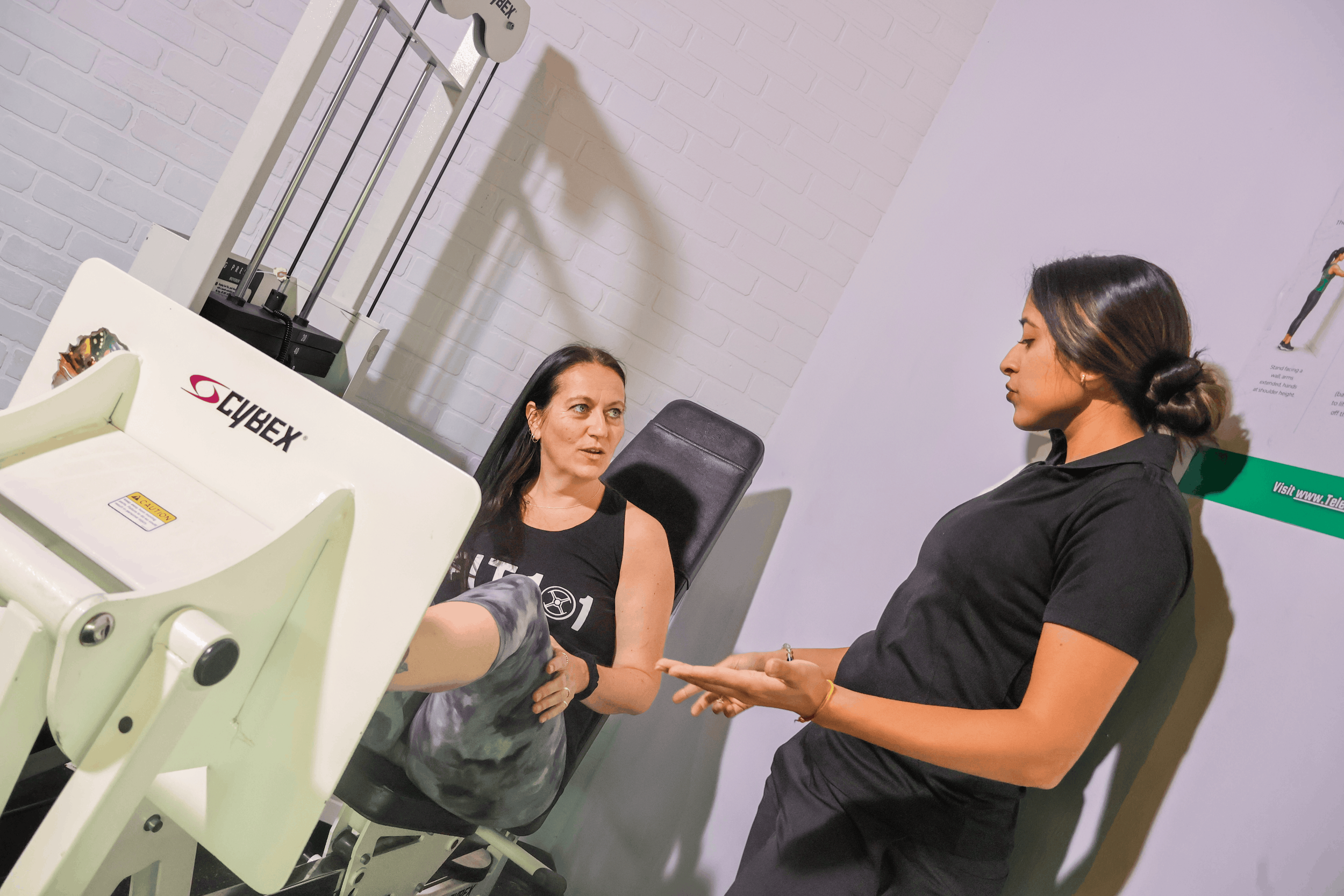 A woman sitting in a chair and a woman in black shirt with similarly long dark hair, standing and talking with her. They are in a room with white brick walls, and there is a CYBEX infant car seat in front of the woman who is seated.