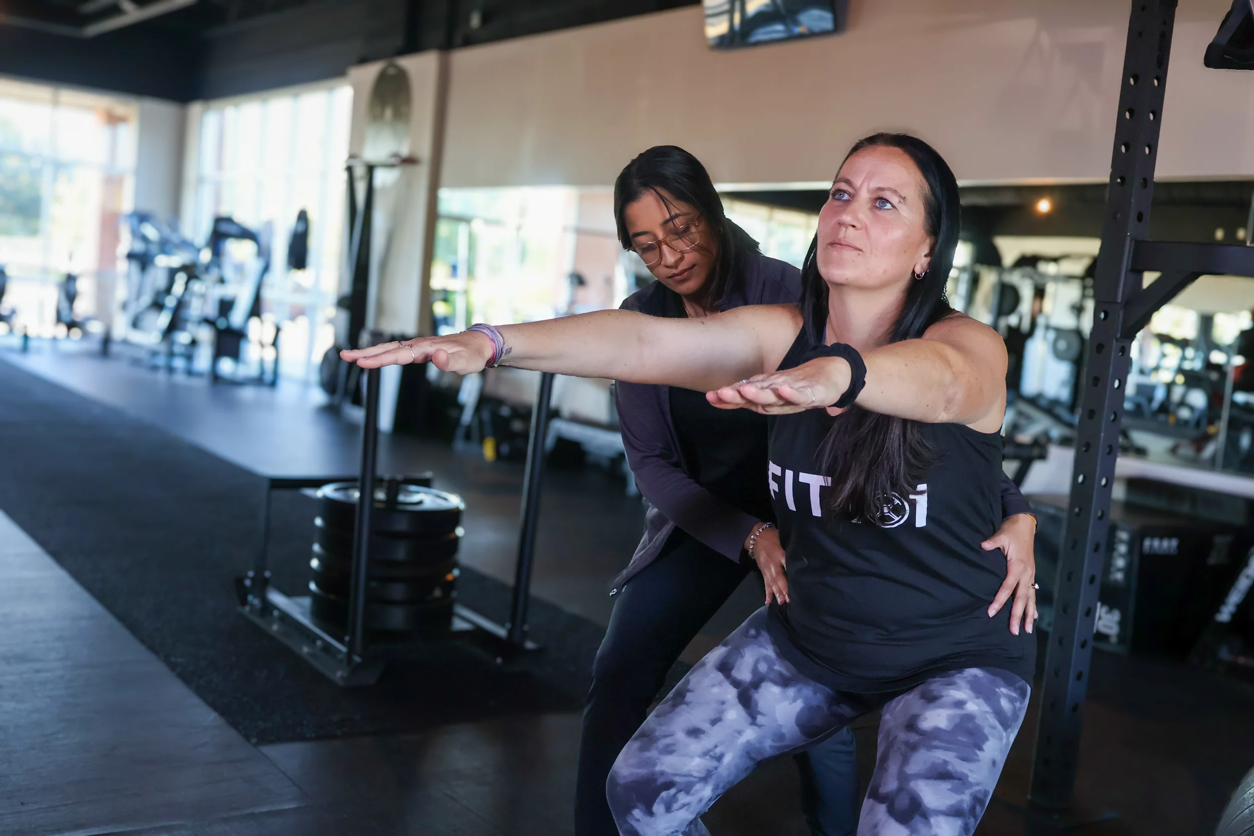 Two women in a gym, one spotting the other as she performs a squat with a barbell, both focused and engaged in workout.