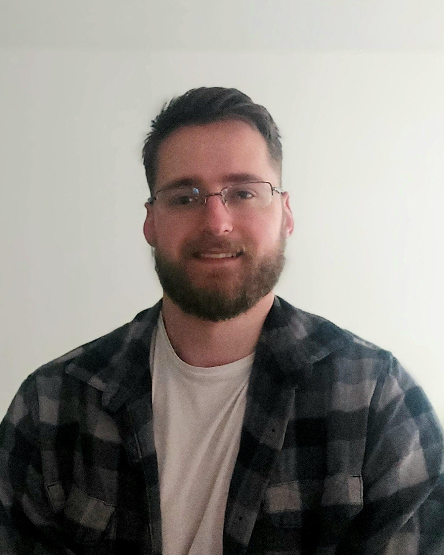 A man with dark hair, beard, glasses, smiling, wearing a plaid shirt over a white t-shirt, against a plain off-white wall.