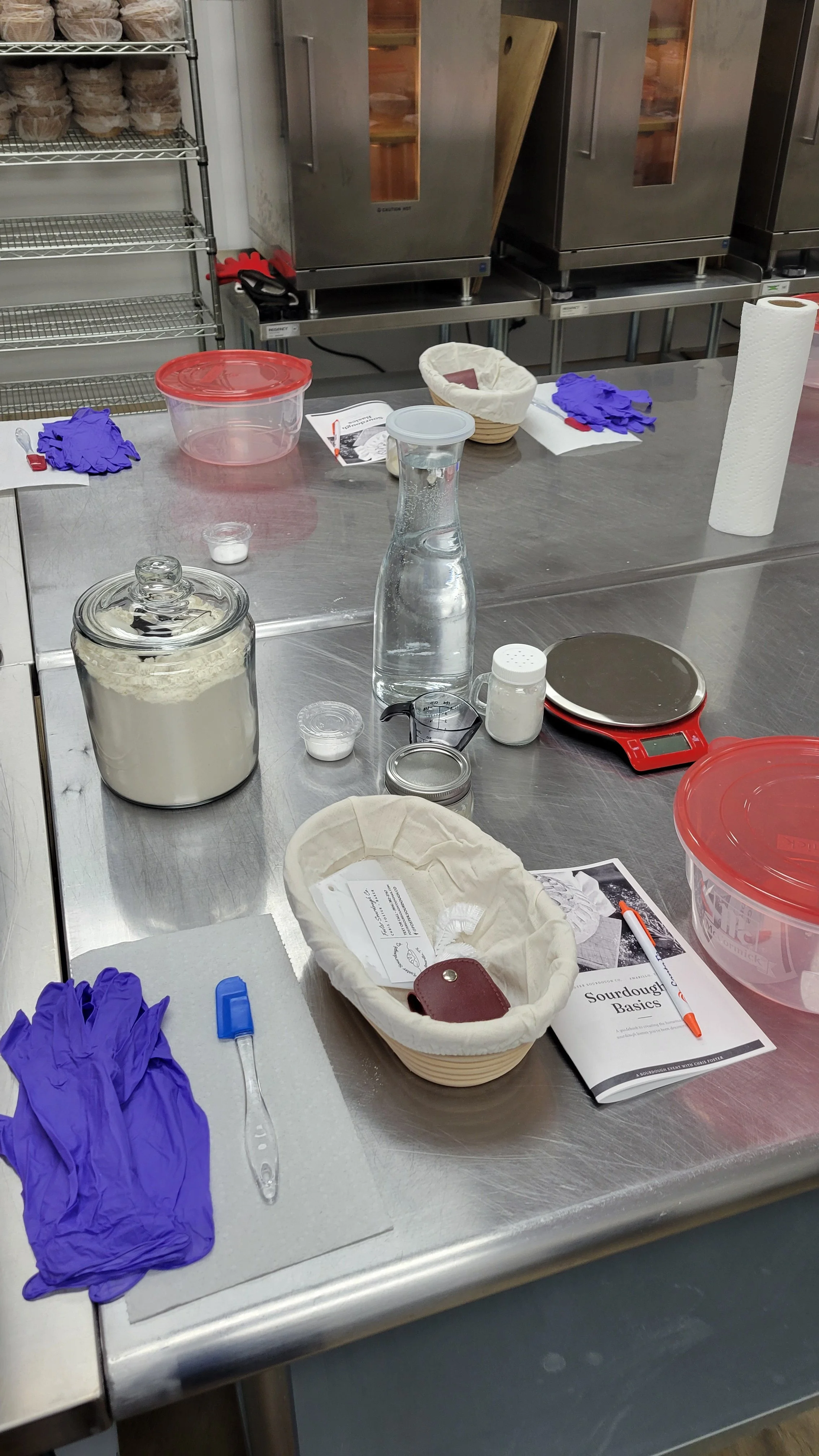 Sourdough class workstation with a jar of starter, water bottle, digital scale, and lined proofing basket.