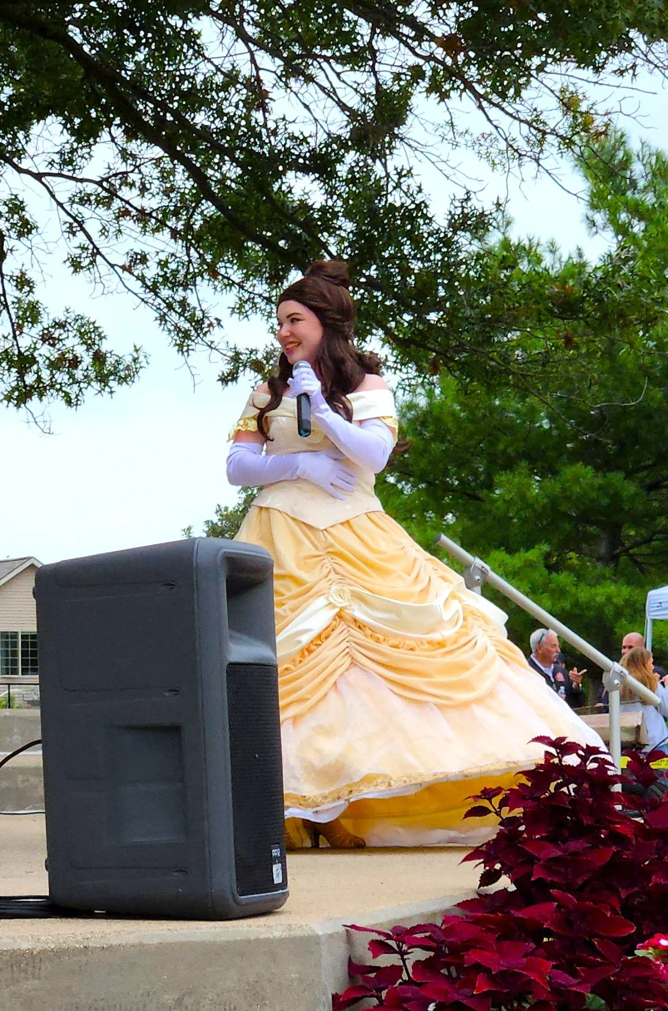 A woman dressed as Belle from Disney's Beauty and the Beast, wearing a yellow gown, standing outdoors on a stage with trees in the background. She is smiling and holding a microphone, with a large speaker in front of her. There are some people visible in the background.