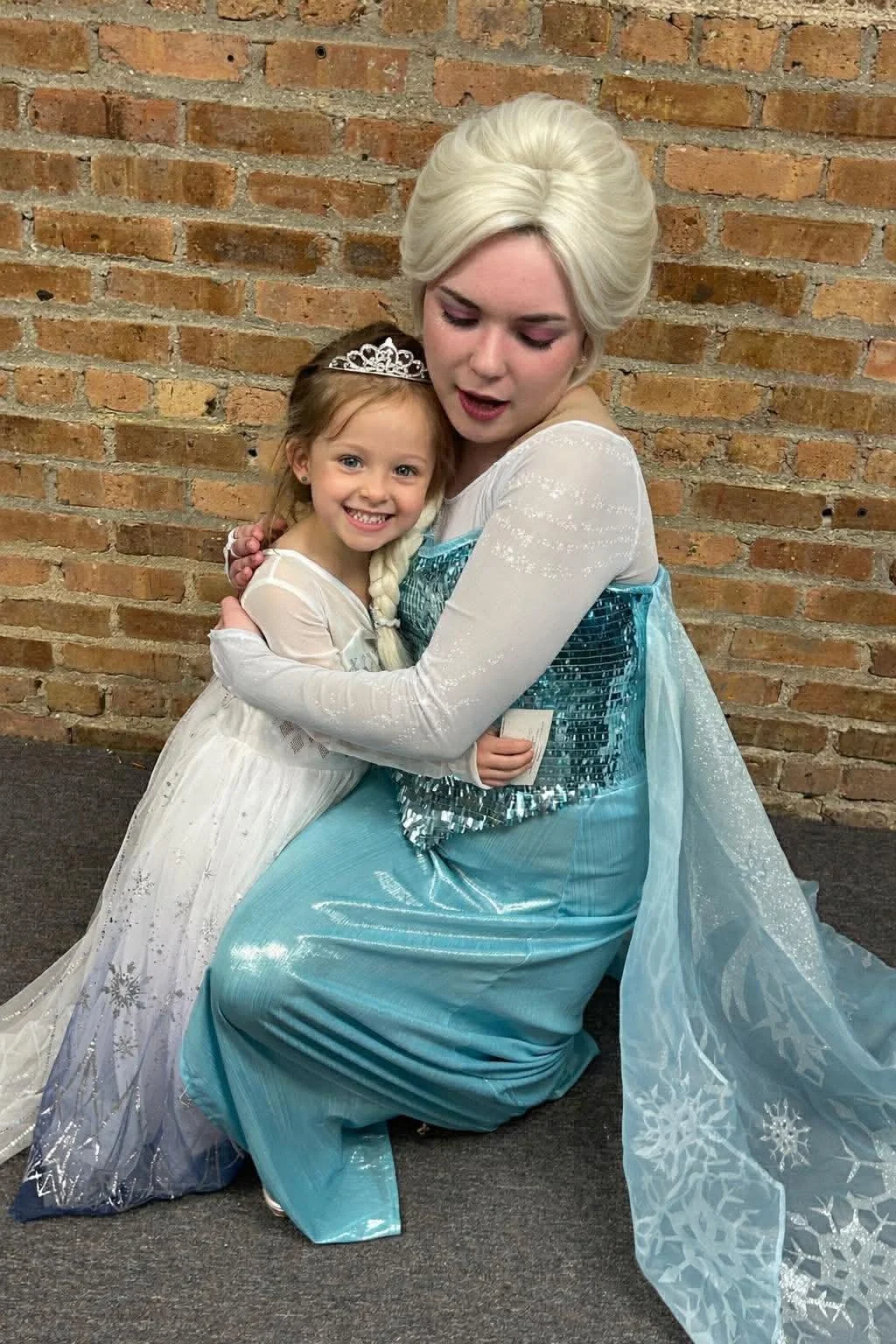 A woman dressed as Elsa from Frozen hugging a young girl in a princess dress, both smiling in front of a brick wall.