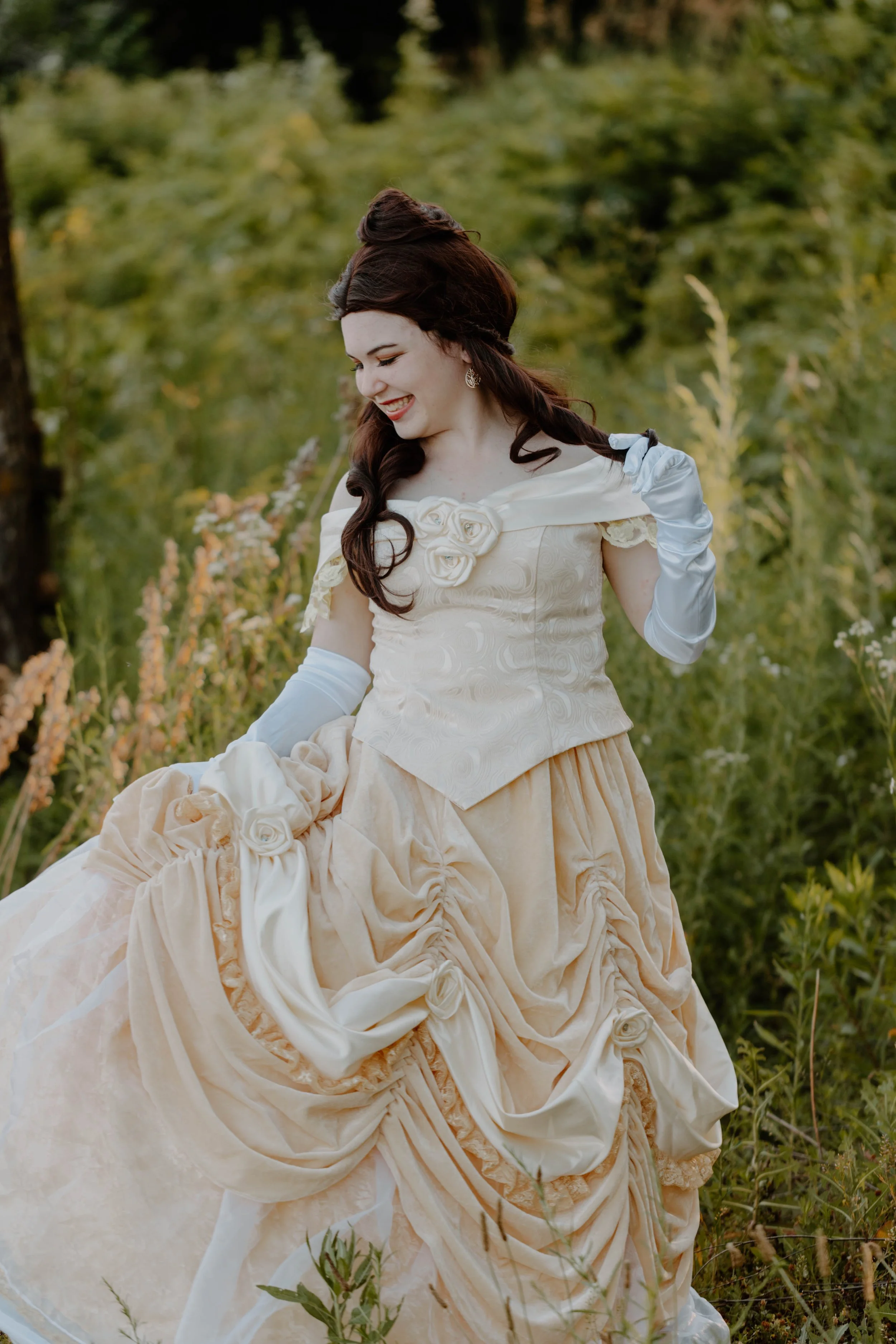 A woman in a vintage cream-colored dress with floral accents, wearing long white gloves, standing outdoors in a natural setting with green trees and tall grass.