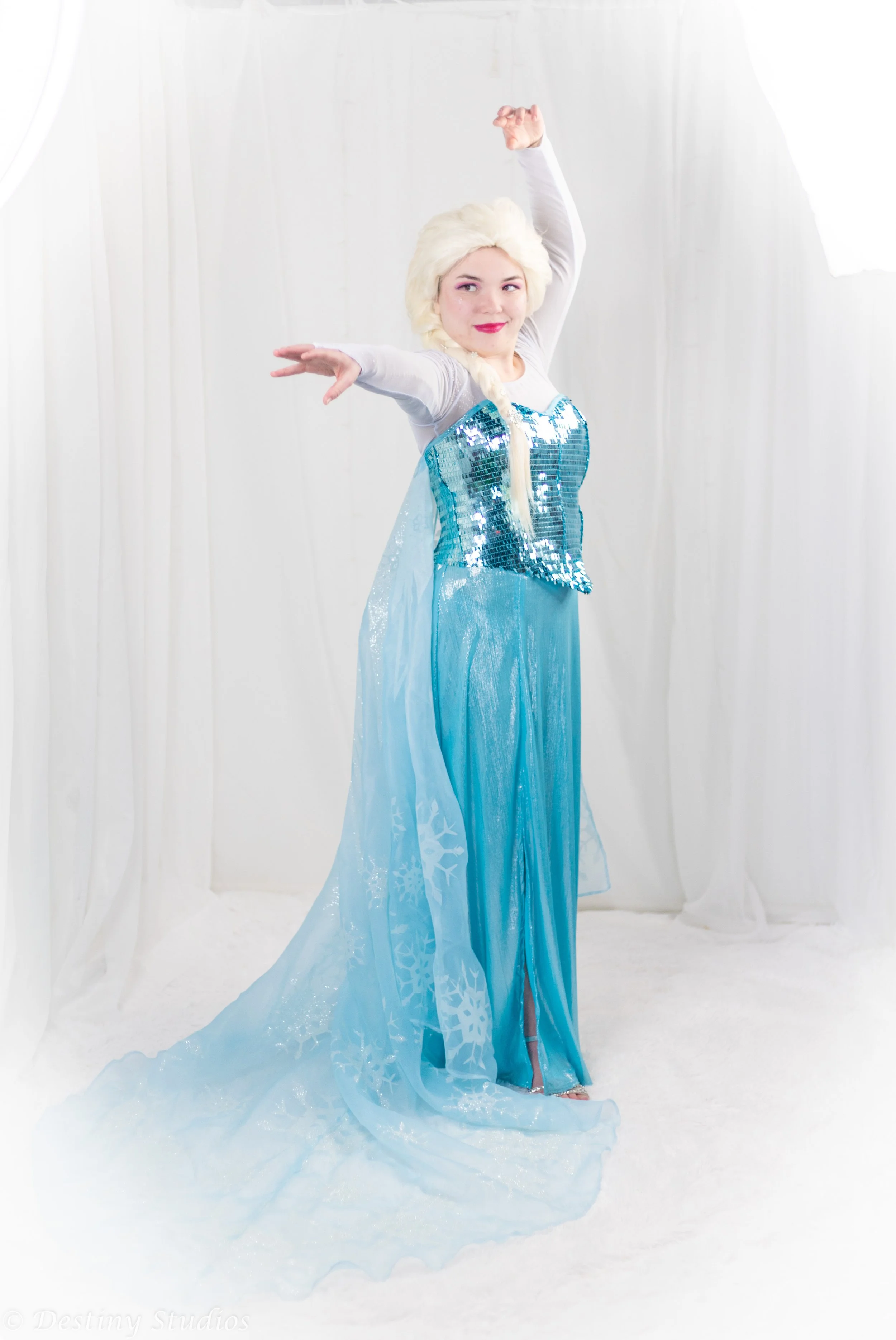 A woman dressed as Elsa from Frozen, standing in a white setting with curtains, wearing a blue dress with snowflake patterns and a sparkly bodice, with platinum blonde hair styled in a braid, smiling and posing with one arm raised.