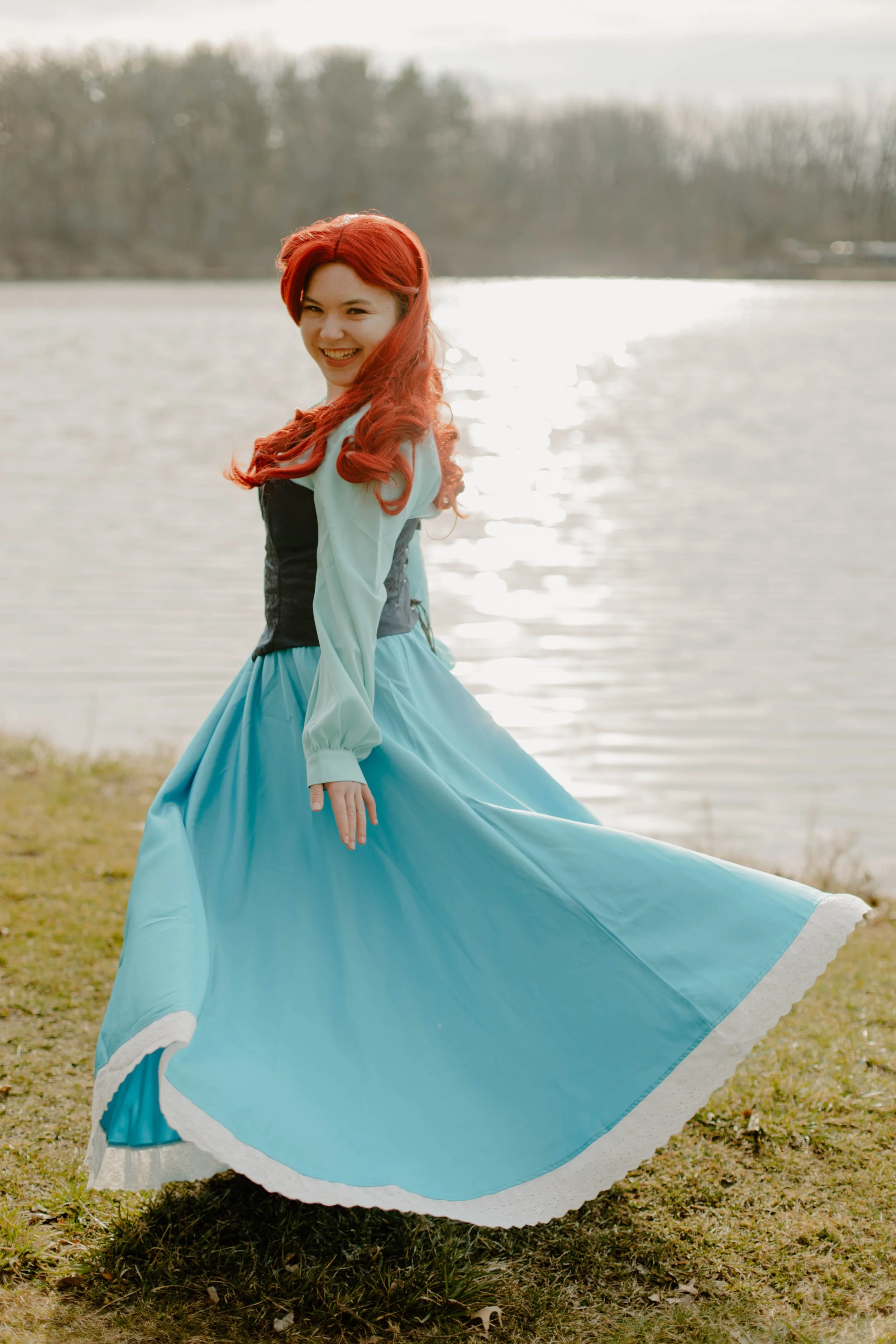 A smiling woman with long red hair in a flowing blue and white dress standing near a body of water with trees in the background.