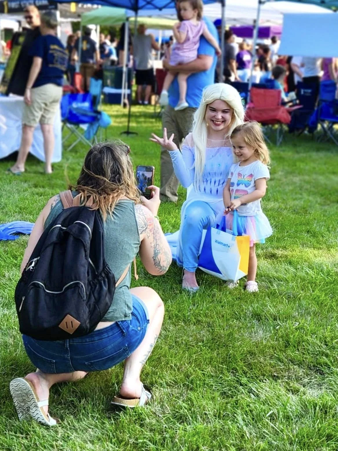 A woman dressed as a princess taking a photo with a young girl, who is holding a shopping bag, at an outdoor event or fair with tents and people in the background.