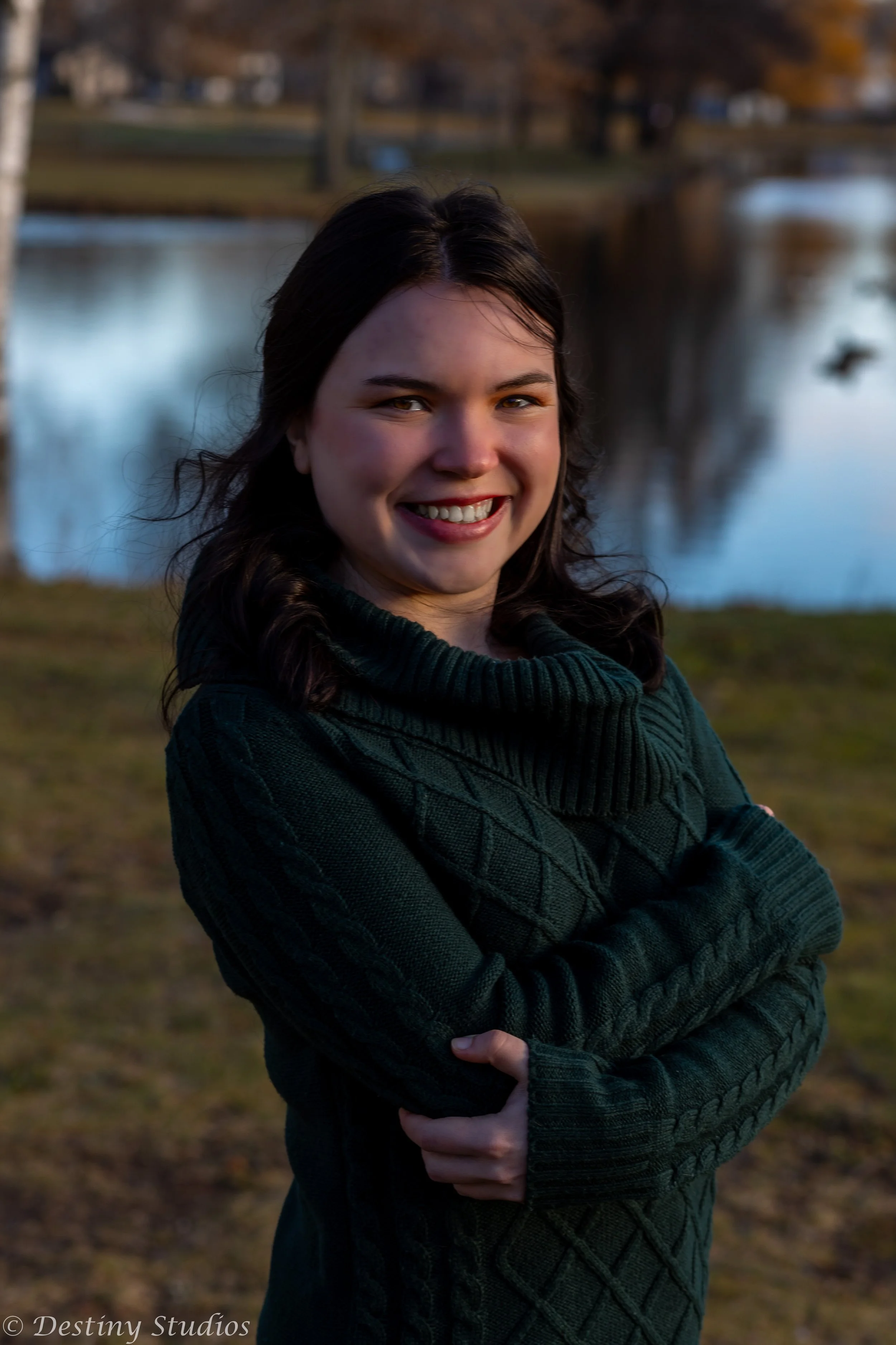 A young woman with dark hair wearing a dark green sweater, smiling, outdoors near a river with autumn trees in the background.