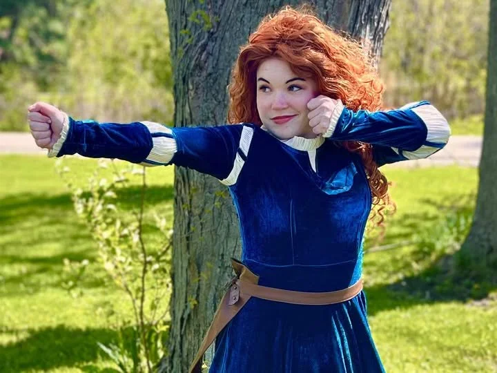 A woman with curly red hair wearing a blue dress and beige belt, standing outdoors near a tree, making a playful punching gesture with both fists, smiling, in a park setting.