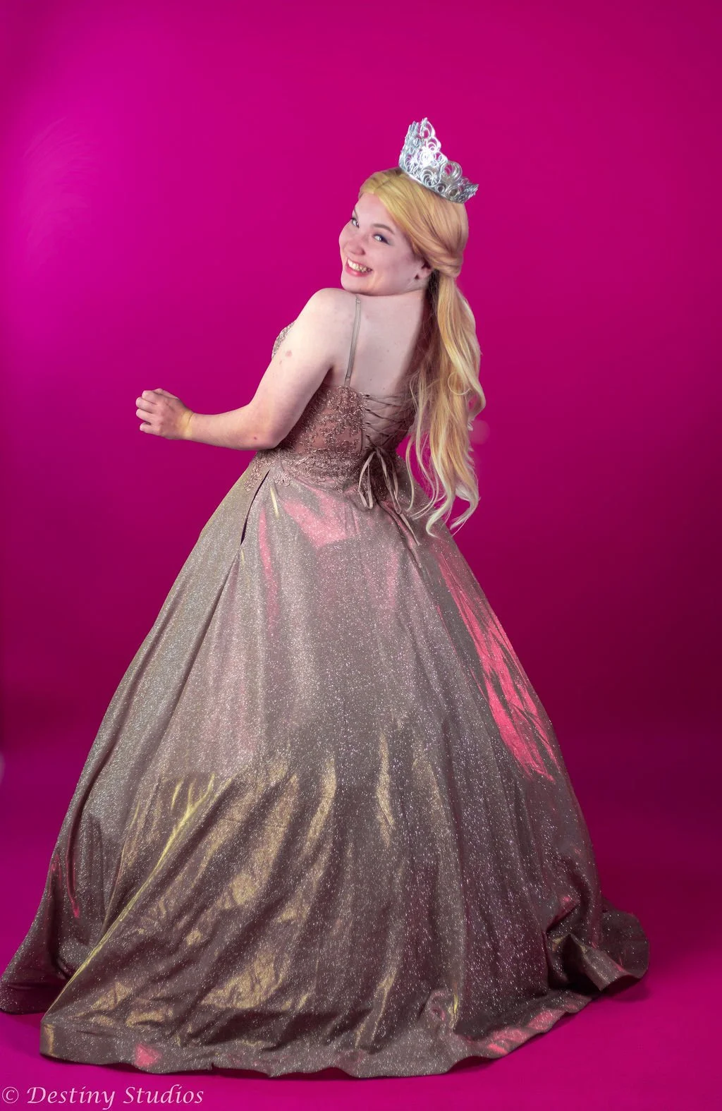 Young woman in a sparkling gold gown with a full skirt, wearing a tiara, standing against a pink backdrop, smiling and turning her head.