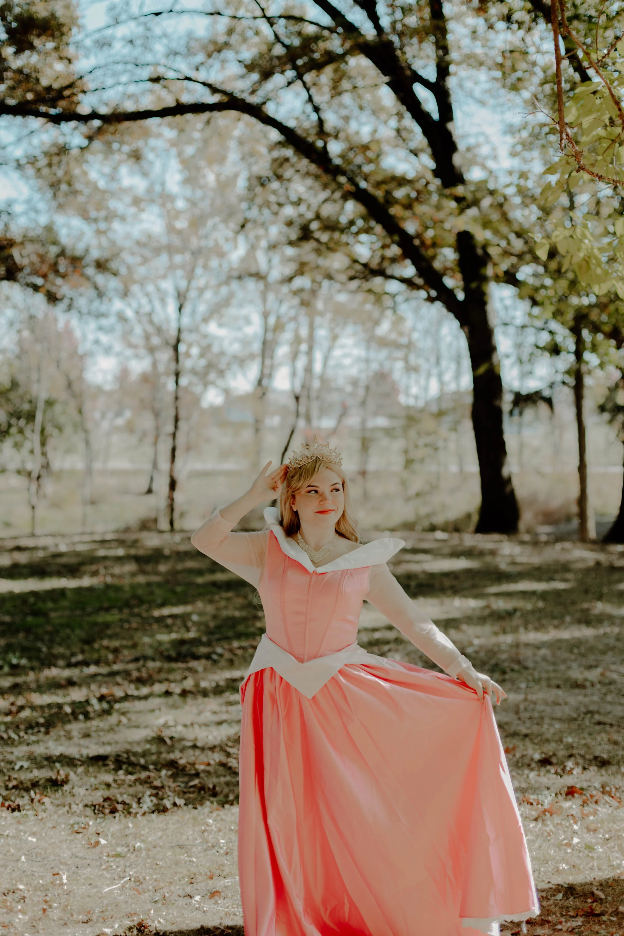 A young woman dressed in a pink gown and princess crown posing outdoors in a park among trees with fallen leaves on the ground.