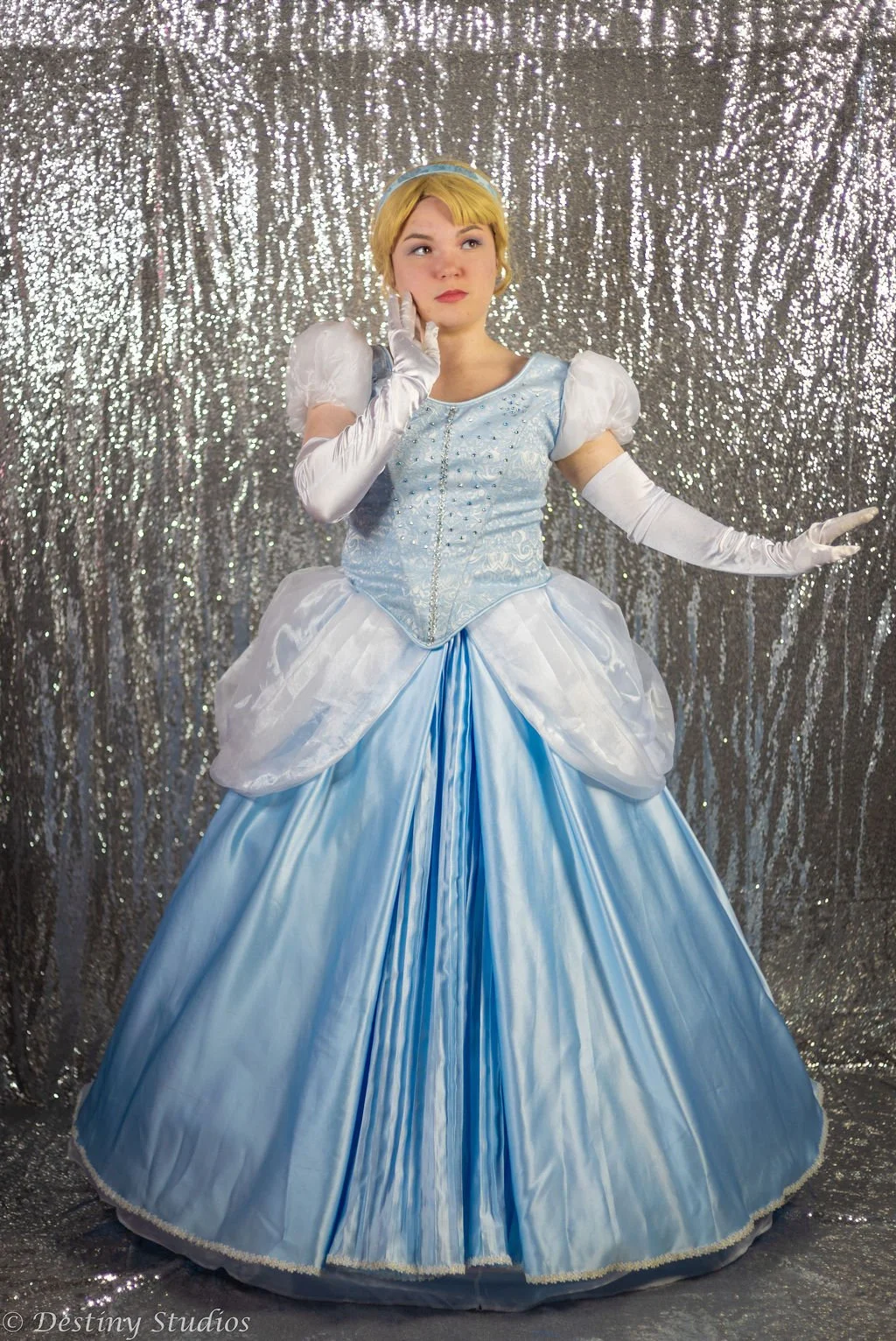 A young woman dressed as Cinderella in a light blue ball gown with puffed sleeves, gloves, and a tiara, standing against a sparkly silver backdrop.