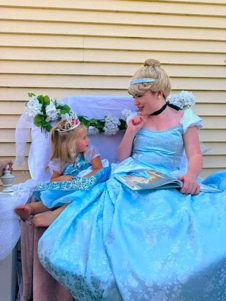A woman dressed as Cinderella, wearing a blue princess gown and a tiara, sits with a young girl dressed as a princess, wearing a tiara and a blue dress. They are outdoors against a yellow wall, with the woman holding a book and the girl looking at her.