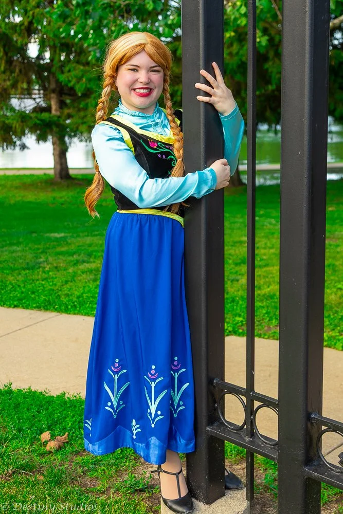A young woman dressed as Anna from Frozen, smiling and hugging a black metal gate, outdoors on a grassy area with trees and a pathway in the background.