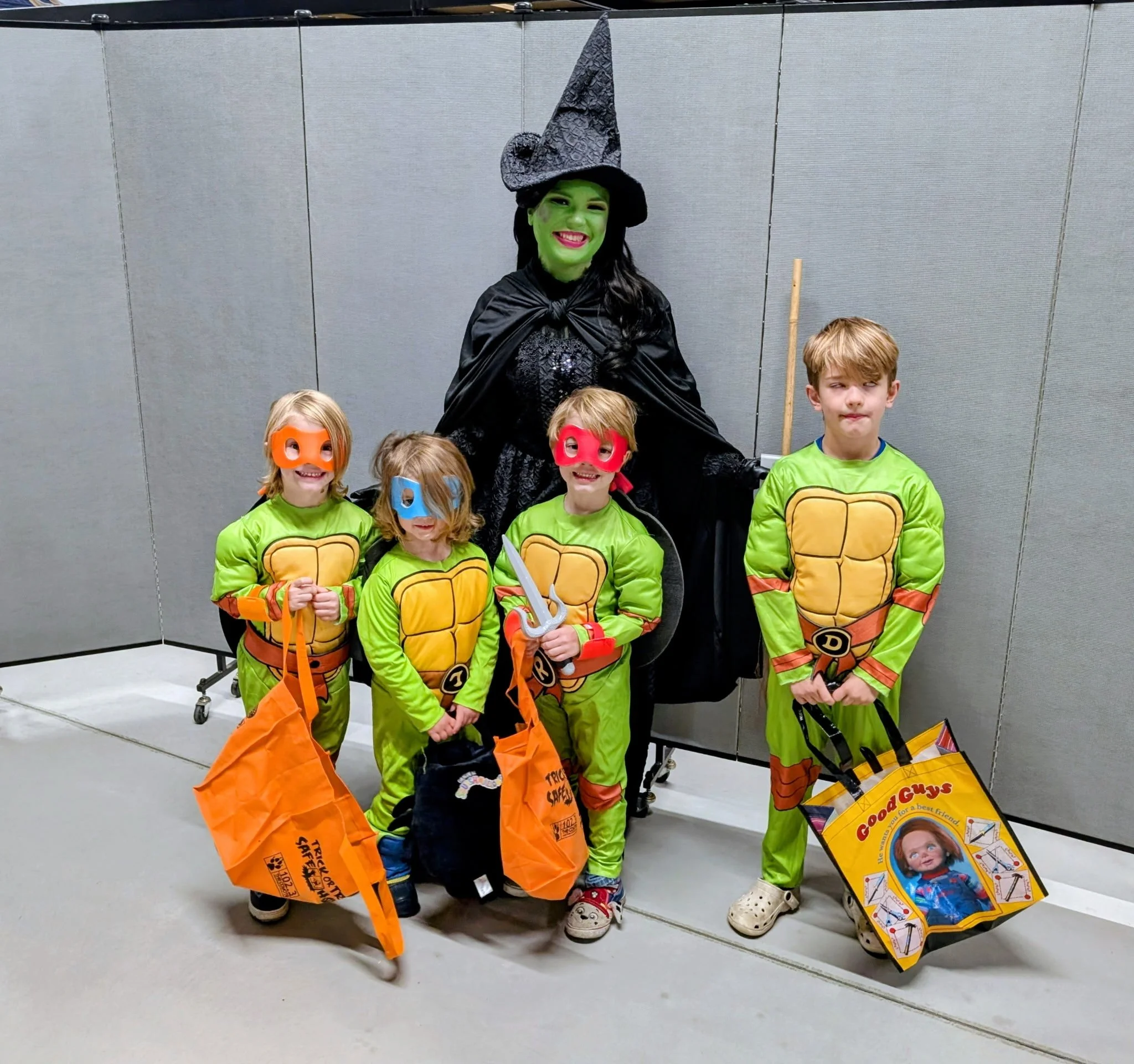 A woman dressed as a witch with green face paint and a tall, pointed witch hat, surrounded by children in Teenage Mutant Ninja Turtles costumes, some wearing masks and holding orange bags, standing in front of a plain gray wall.