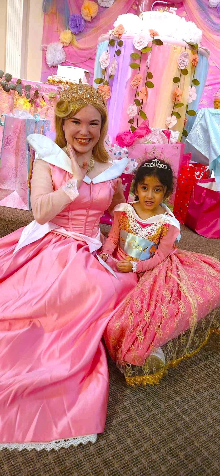 A woman and a young girl dressed as princesses, sitting on the floor at a birthday party with pink decorations, gift bags, and flowers.