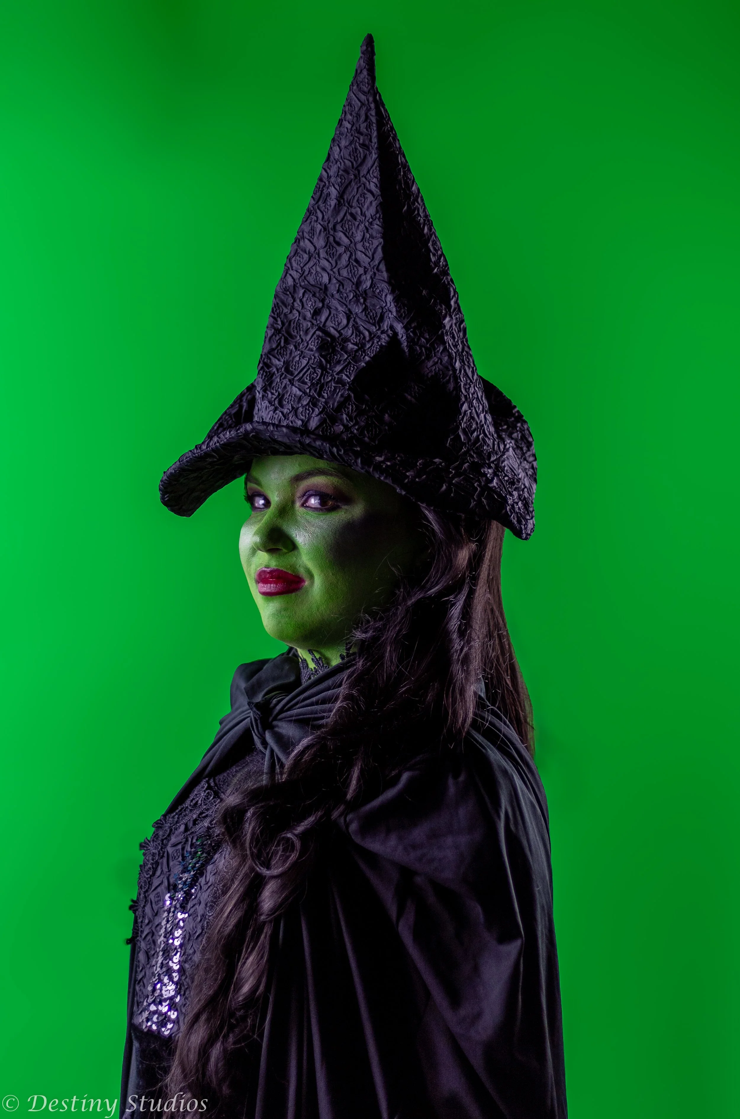Woman dressed as a witch with a tall, textured black hat and green face makeup, wearing a black cloak against a green background.