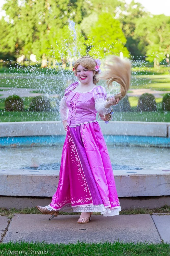 Woman in pink princess dress standing in front of a fountain, smiling, with water splashing behind her, surrounded by green trees.
