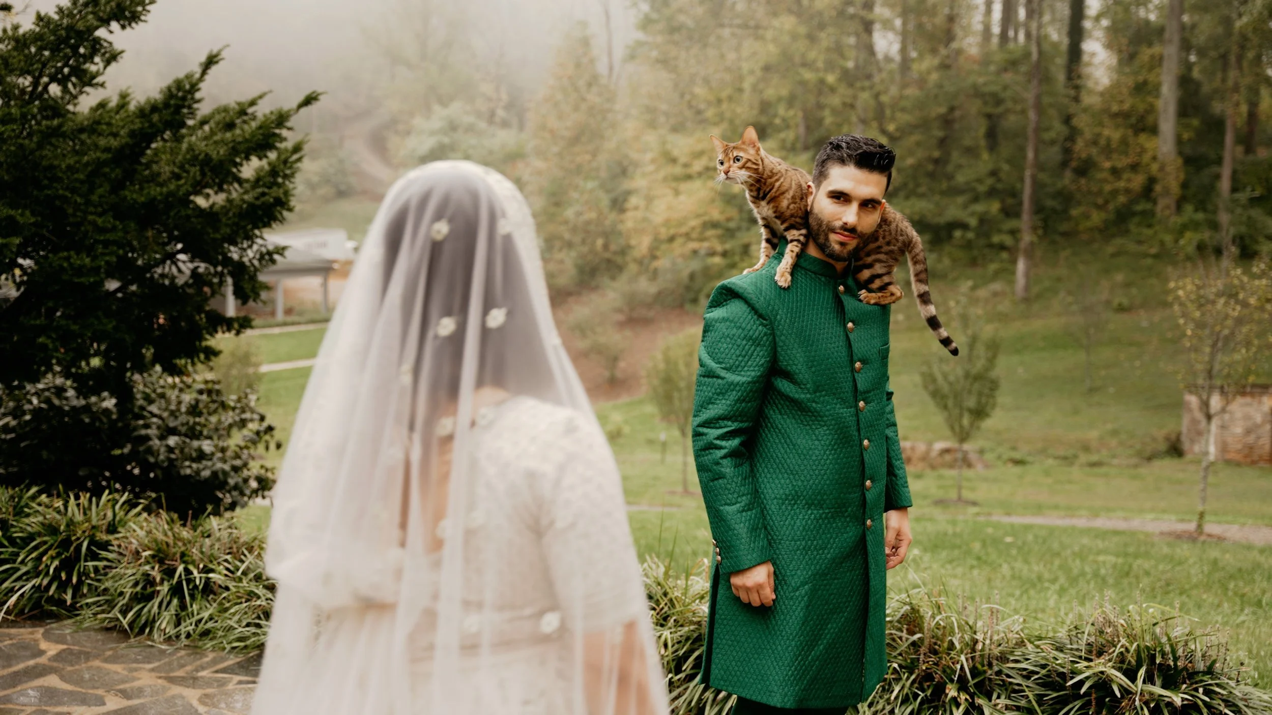 Un hombre con barba y, vestido con un abrigo verde, está frente a una mujer con un velo de novia, en un entorno al aire libre con árboles y vegetación, y detrás de él hay un gato atigrado sobre su hombro.