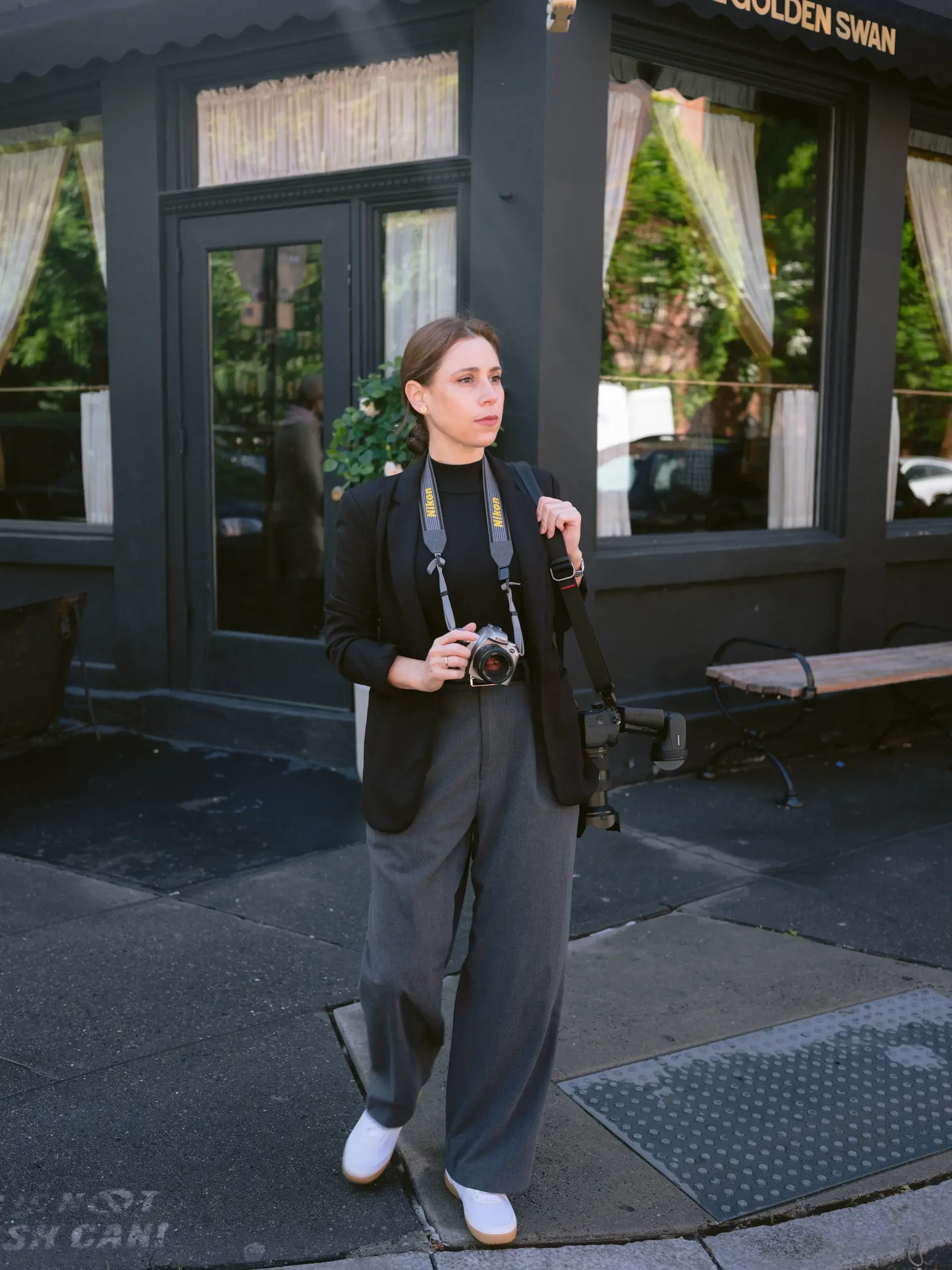 Mujer joven con cabello castaño, vestida con blazer negro y pantalones grises, con un libro y una cámara en las manos, en la acera frente a un edificio con ventanas con cortinas blancas y un letrero que dice 'Golden Swan'.