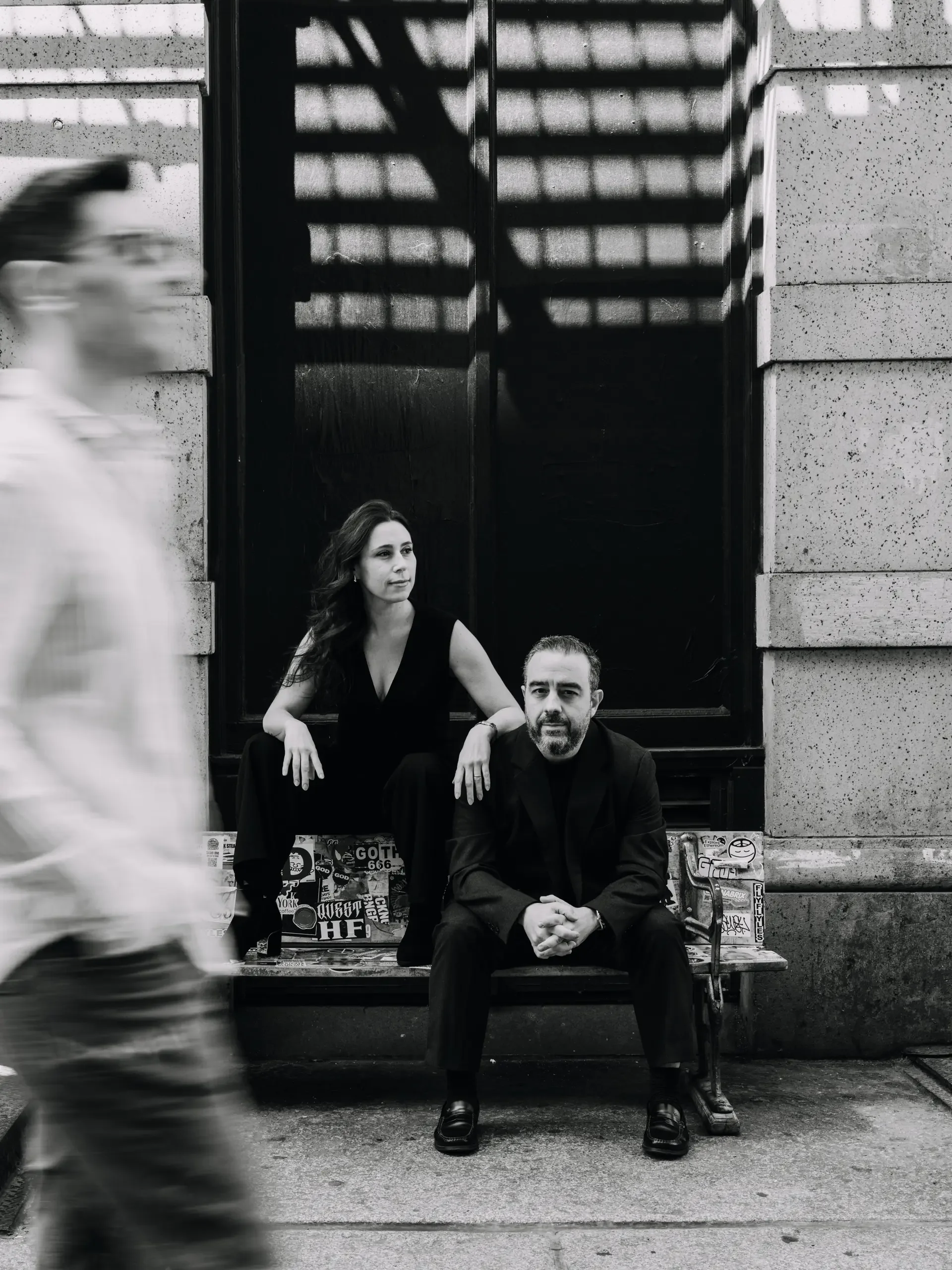 Dos personas sentadas en una banca en la acera frente a un edificio con paredes de piedra. Una mujer con cabello rizado y ropa negra, y un hombre con barba, cabello corto y ropa negra. Una persona con camiseta con rayas pasa en primer plano, borroso.