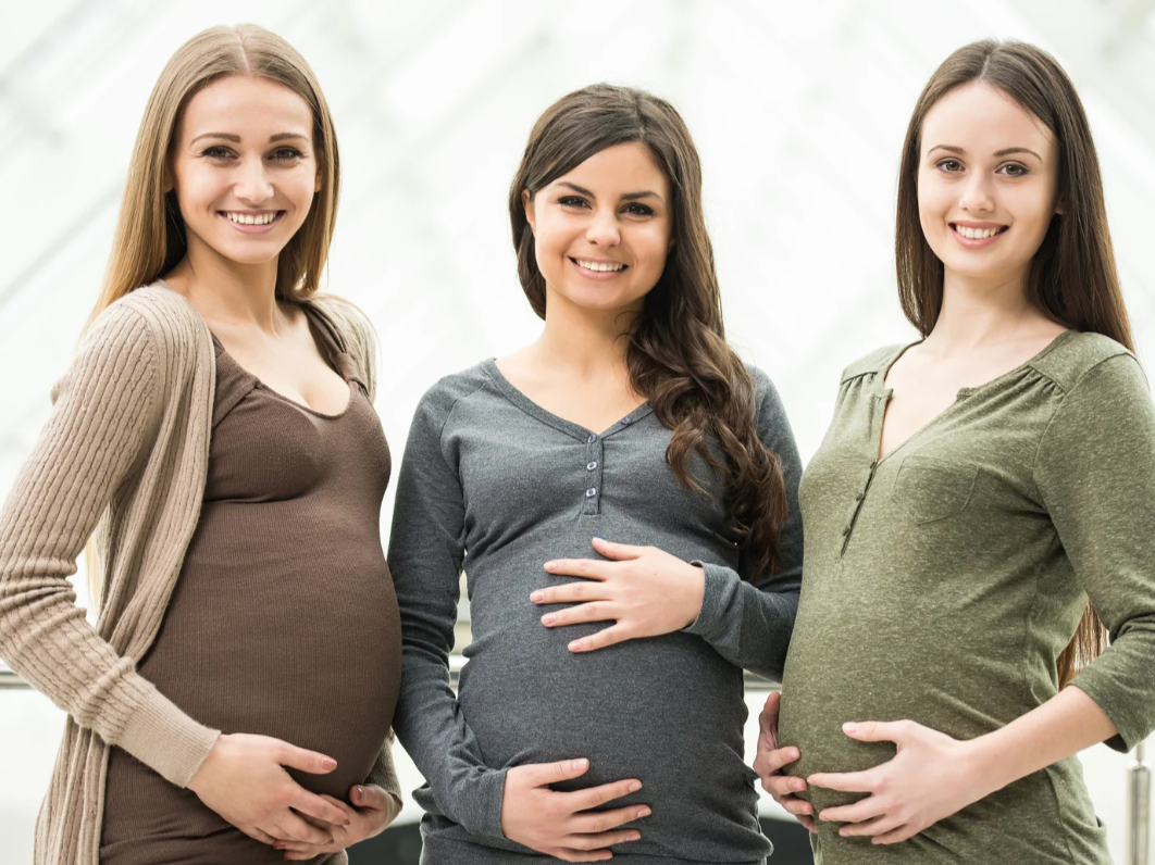 Three pregnant women standing together indoors, smiling, with their hands on their bellies.