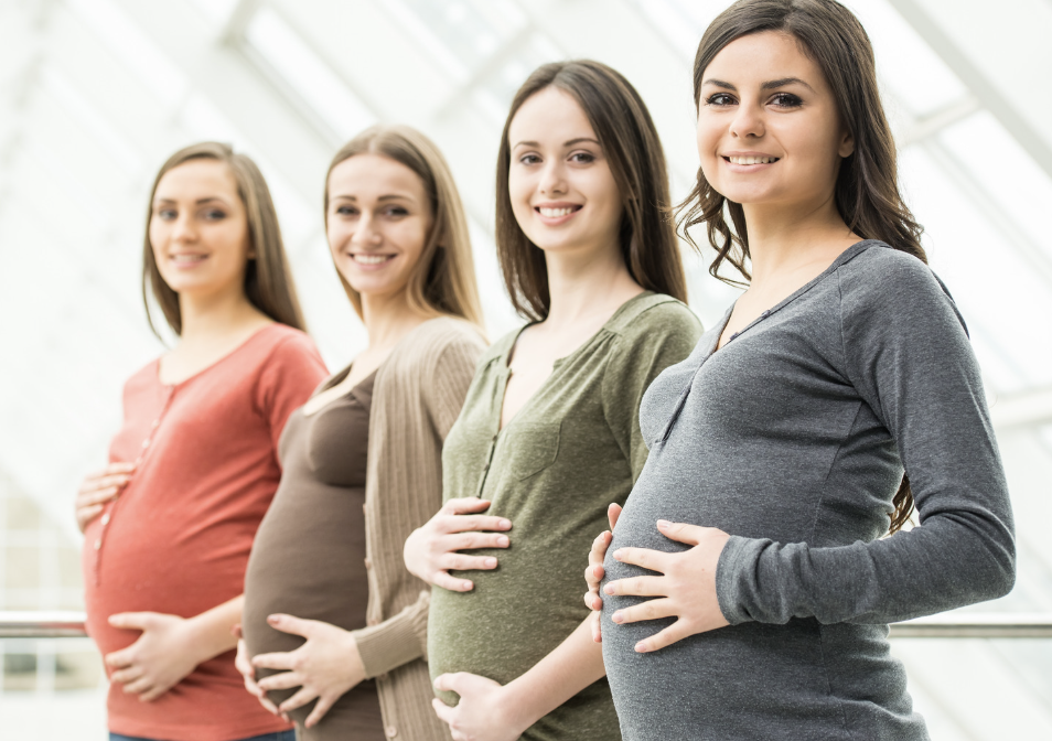 Four pregnant women standing in a row indoors, each holding her belly, smiling at the camera.