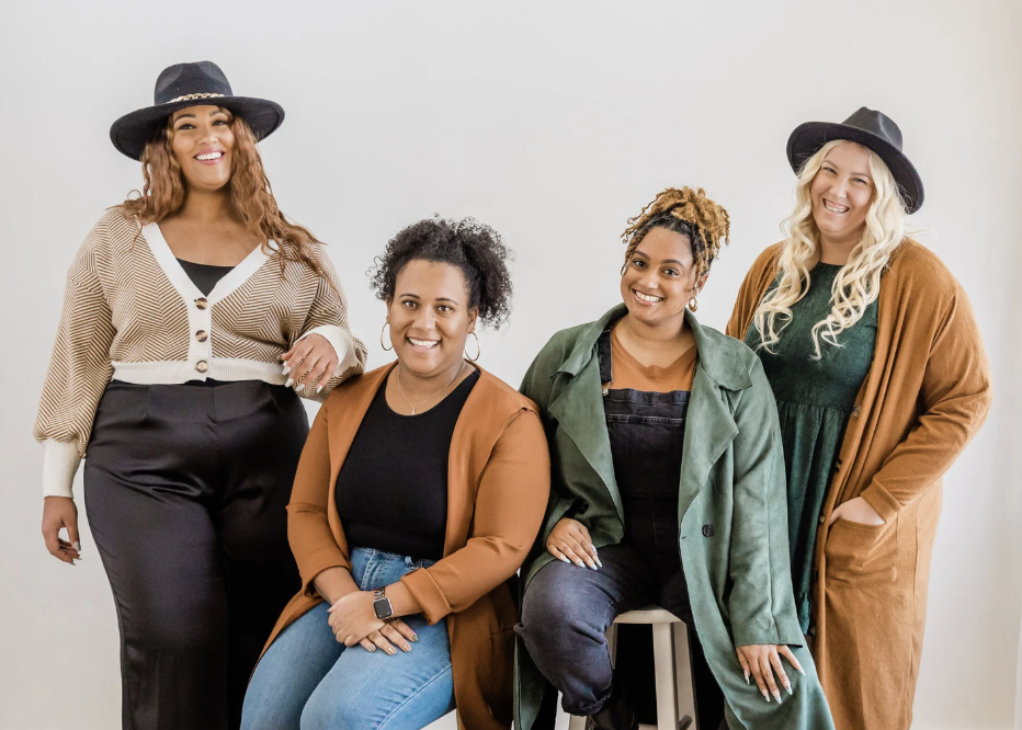 Four women smiling and posing together in casual stylish clothes and hats against a plain white background.