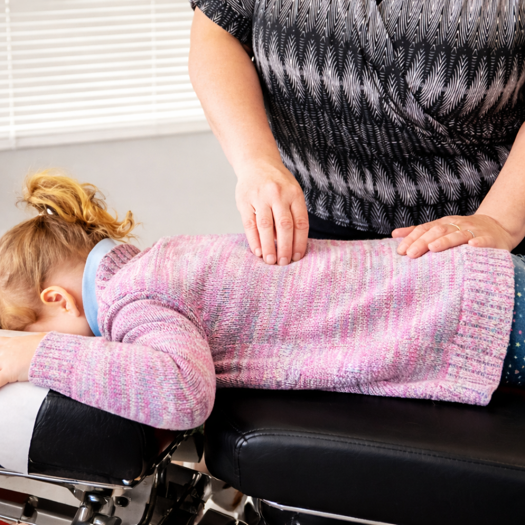 A toddler receiving a back adjustment from a female chiropractorwhile lying face down on a chiropractic table, wearing a pink and purple sweater.