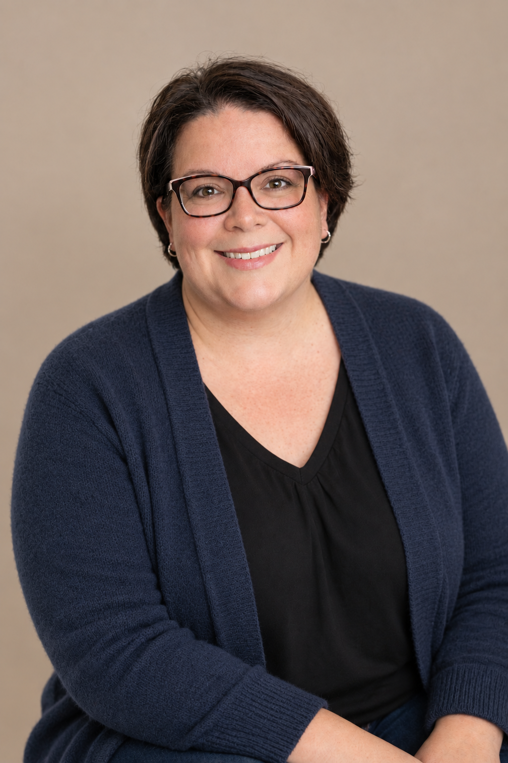 Portrait of a smiling woman with short dark hair, wearing glasses, a black top, and a navy blue cardigan against a neutral background.