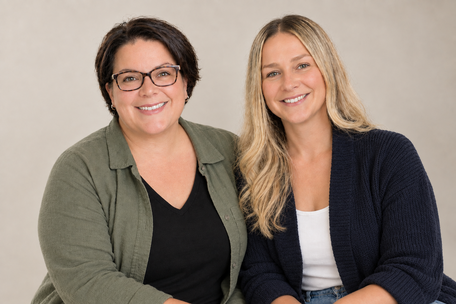 Two women smiling and sitting closely together against a neutral background.