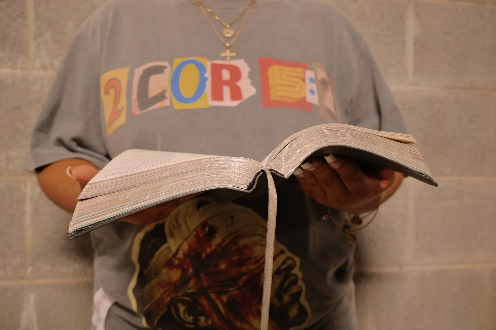 Person standing against a brick wall, wearing a gray T-shirt with colorful, cut-out style letters spelling '2COR 5:7' and holding an open bible.