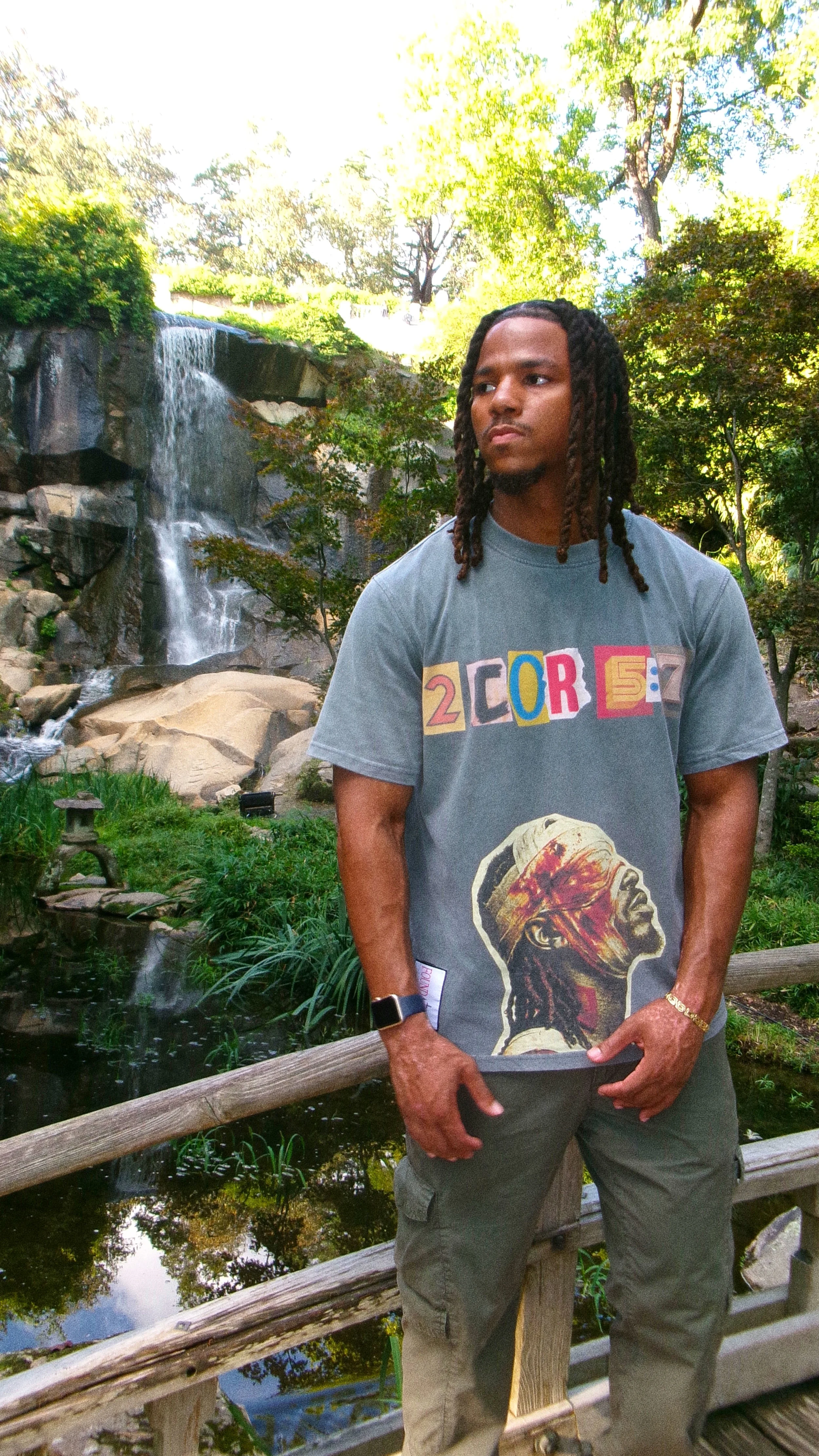 A man with long dreadlocks wearing a graphic t-shirt and standing on a wooden bridge in front of a waterfall and lush green trees.