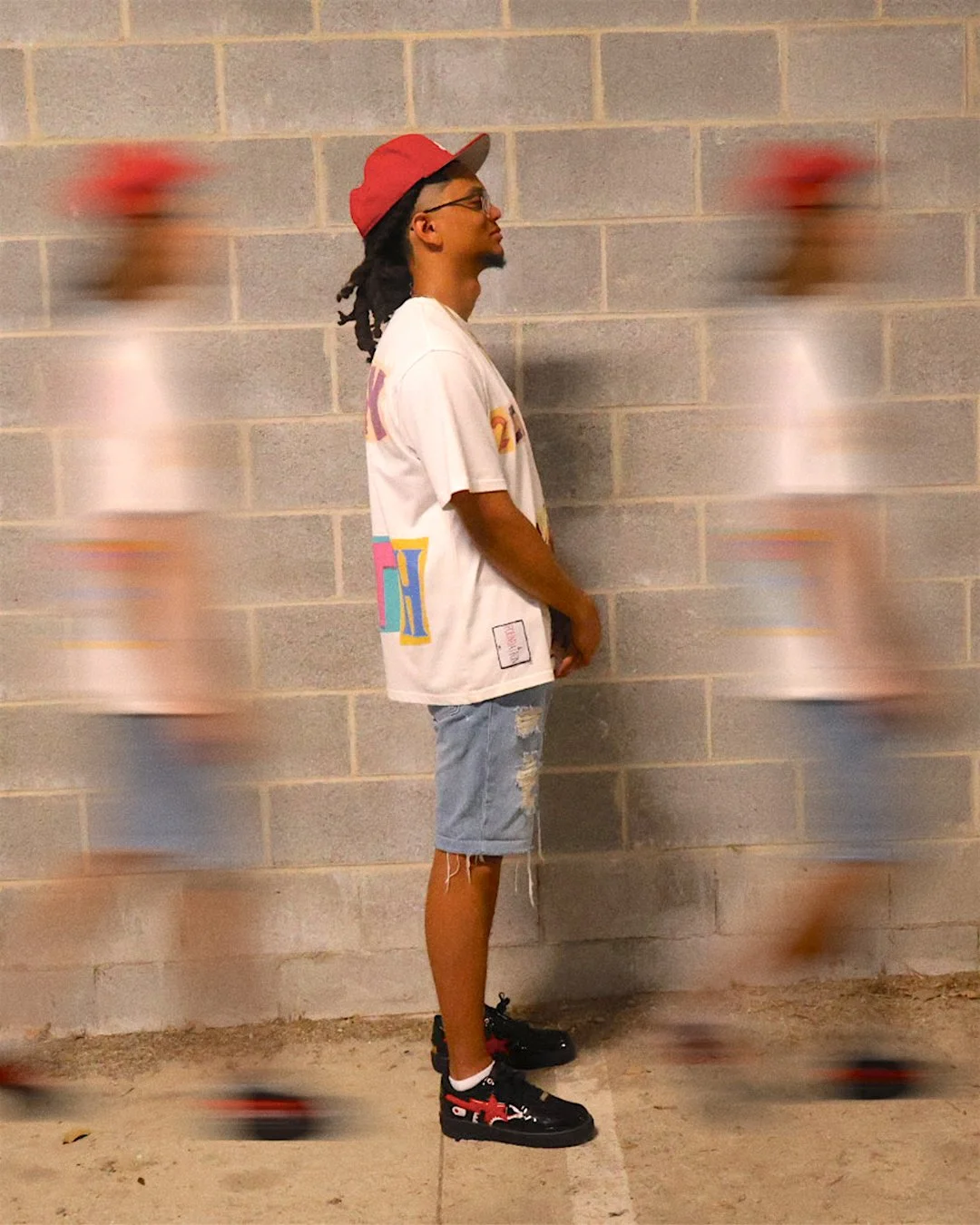 A young man with dreadlocks, glasses, and a red baseball cap stands in profile against a concrete block wall. He wears a white graphic t-shirt, ripped jean shorts, and black sneakers with red accents, while skateboarders are blurred around him.