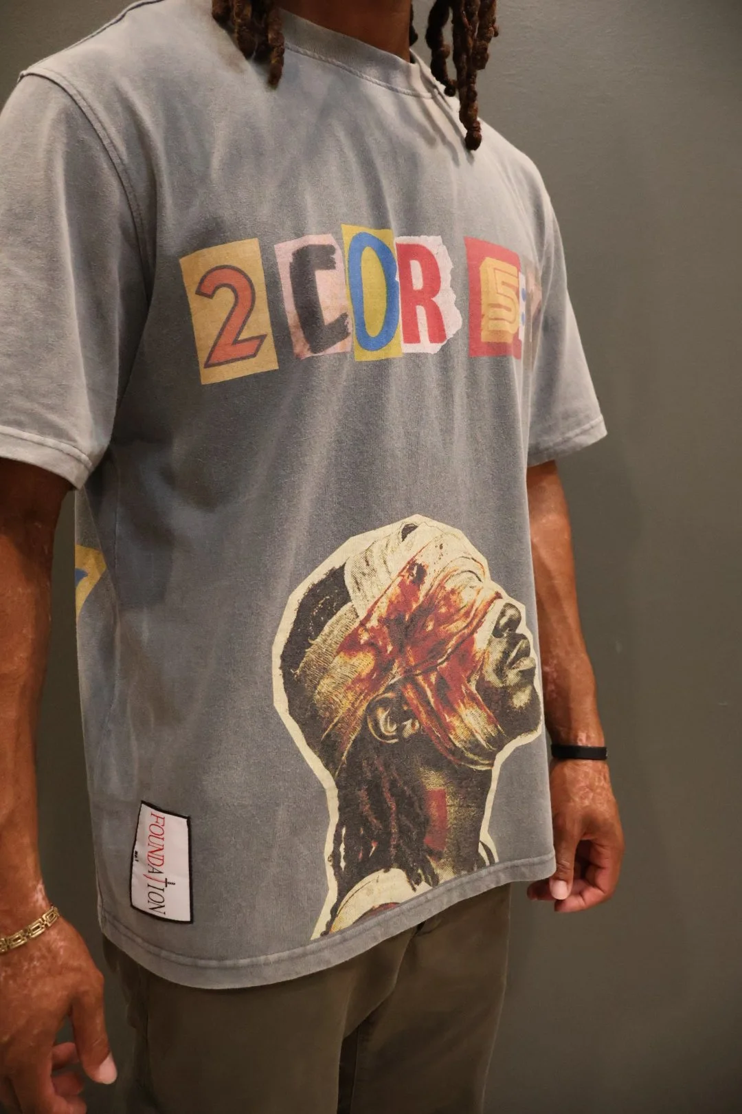 A person wearing a gray T-shirt with colorful letters spelling "2COR5:7" and an illustration of an African-American man with a cloth covering his eyes, a headwrap, and dreadlocks.