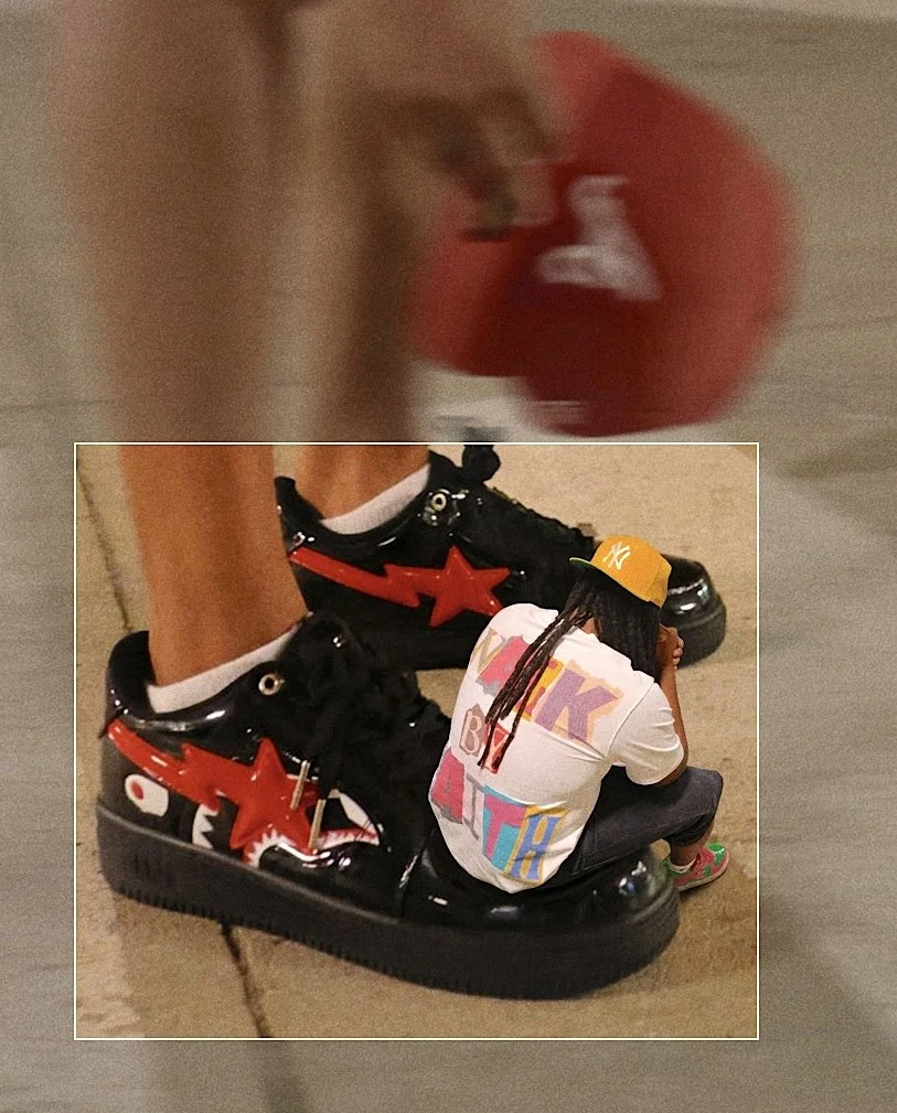 A person wearing black sneakers with red star designs and yellow eyelets, with a small figure of a person in a white T-shirt, yellow hat, and colorful shorts sitting on the sneakers.