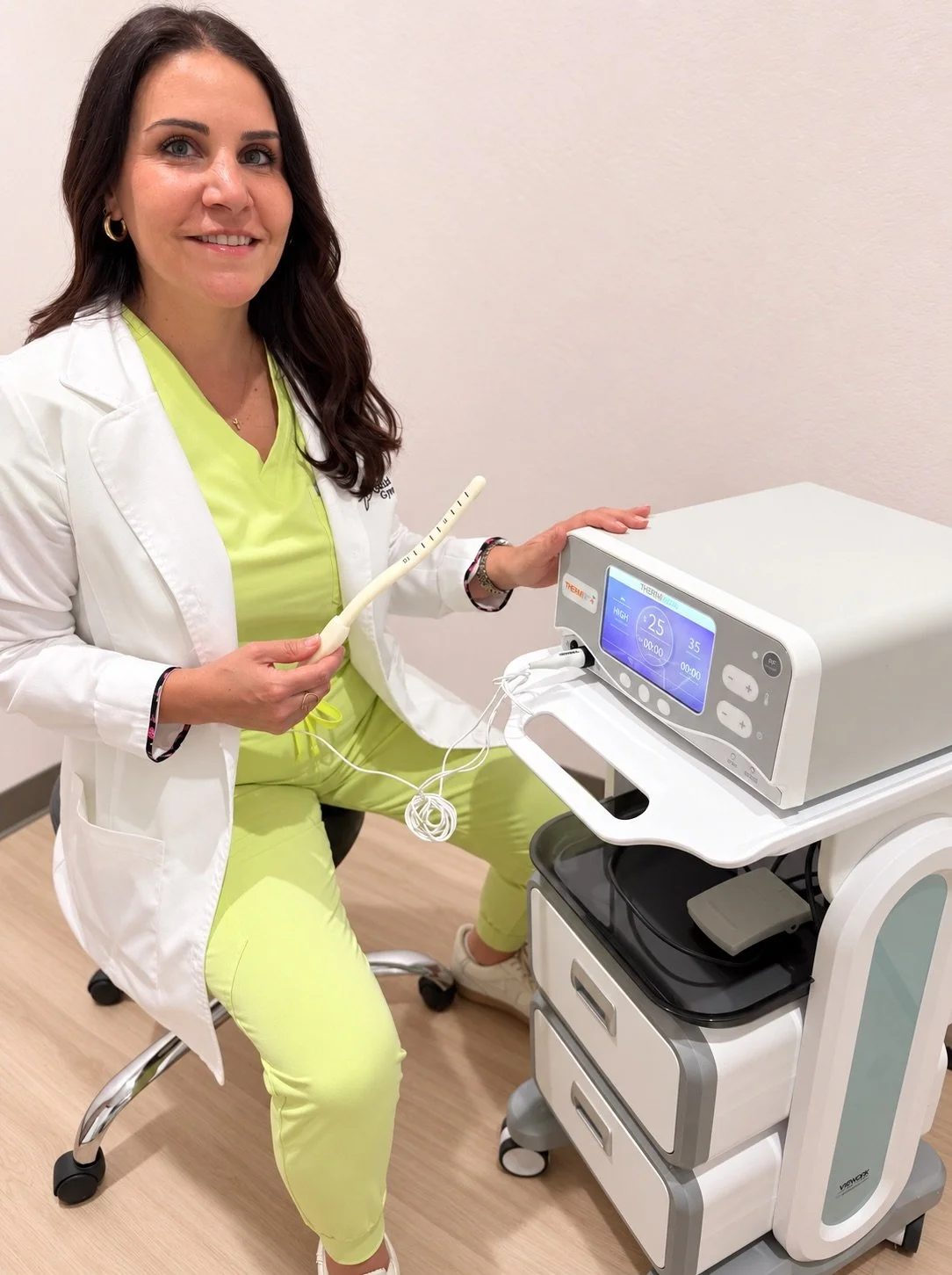 A healthcare professional in a white coat and lime green scrubs sitting on a rolling stool, holding a medical probe, next to a medical device digital console, with a plain white wall in the background.