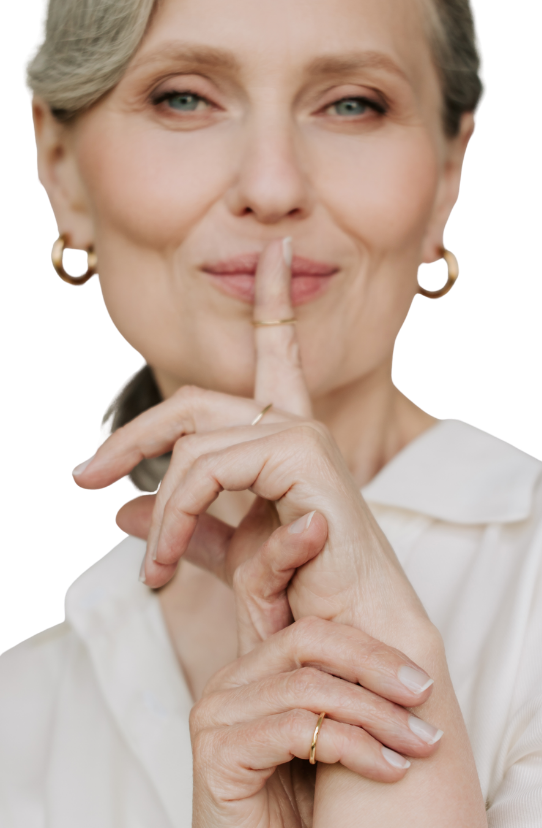 A mature woman with short, gray hair making a 'shh' gesture by placing her index finger on her lips, smiling softly, wearing a white blouse and small hoop earrings.