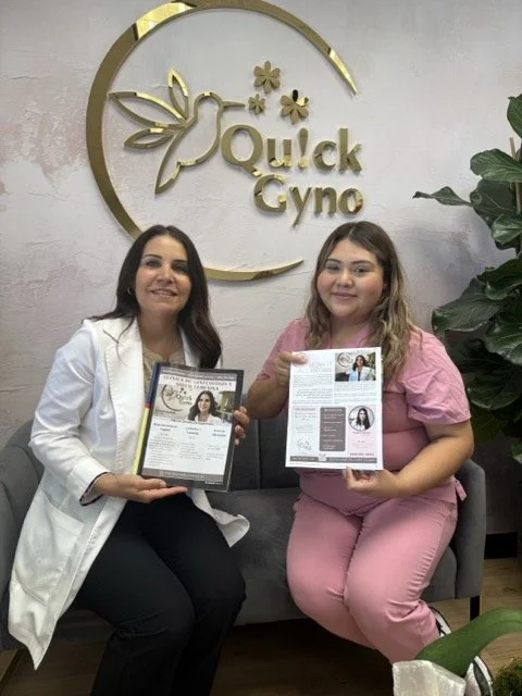 Two women sitting on a gray couch, holding informational brochures, in front of a wall with a gold logo that says "Quick Gyno" and has a hummingbird and flower design, with green plants nearby.