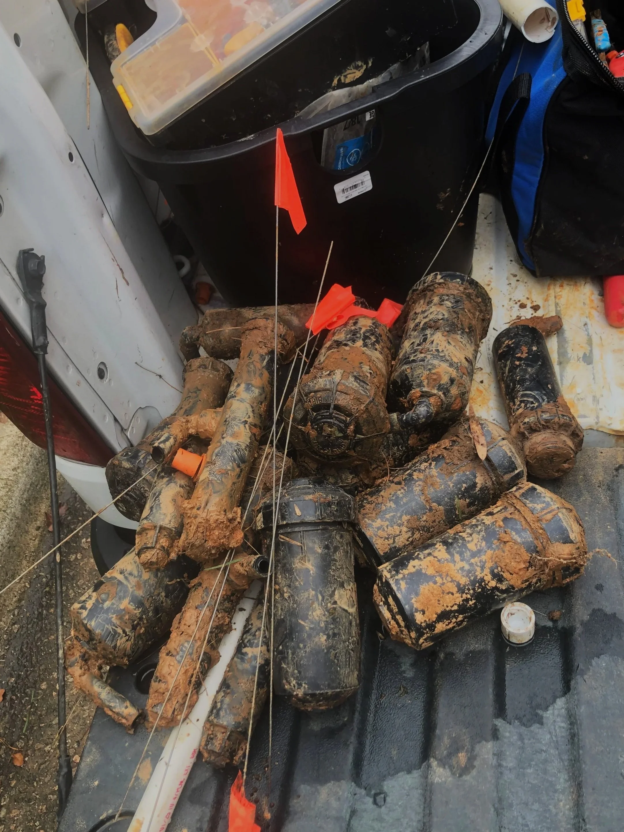 A collection of rusted and muddy pipes and parts on a black surface next to a white truck. There are orange flags and a black and blue bag in the background, along with a muddy white pipe and a white plastic container.