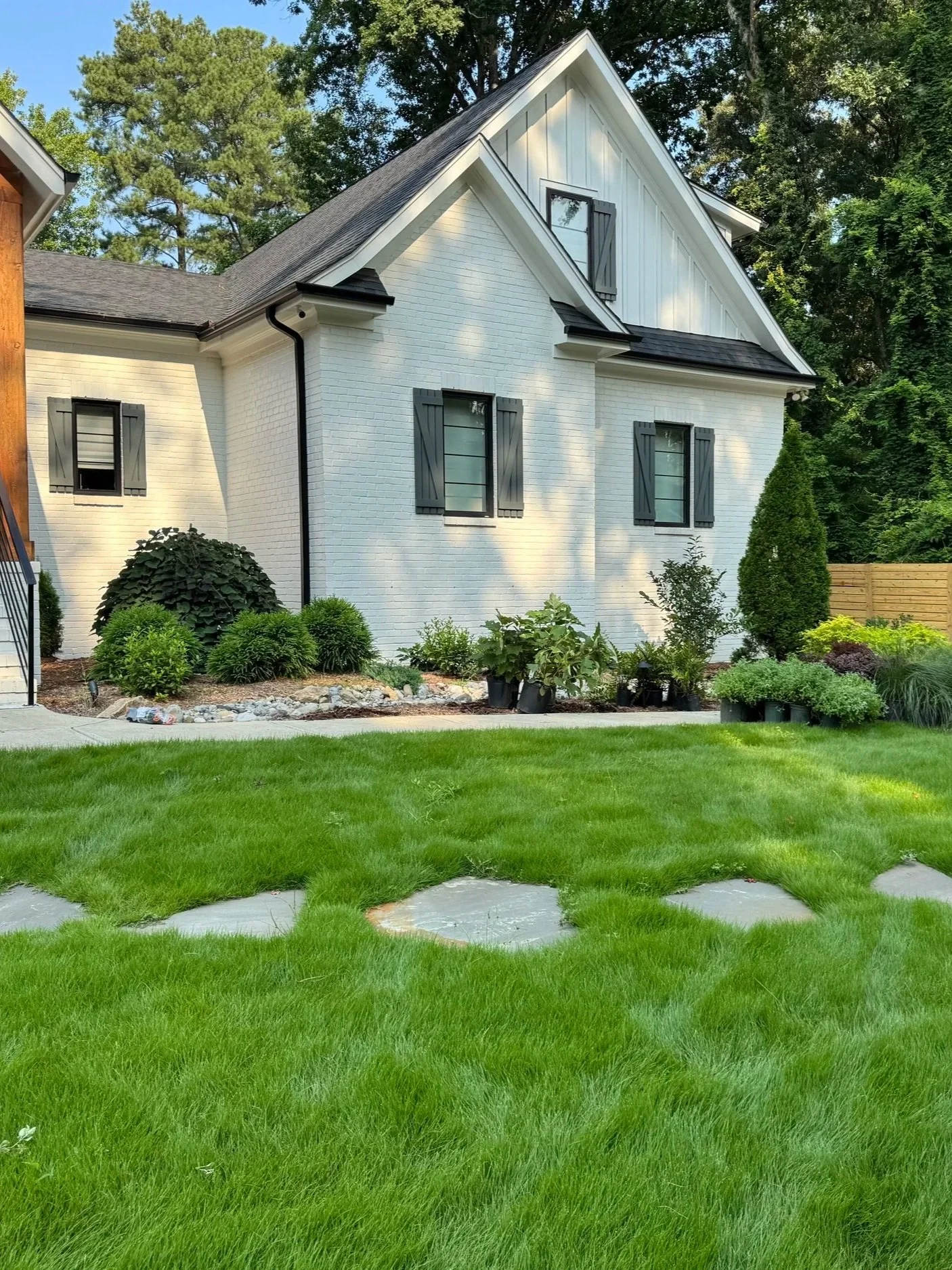 A white house with black window shutters, surrounded by green trees and a well-maintained lawn with stepping stones.