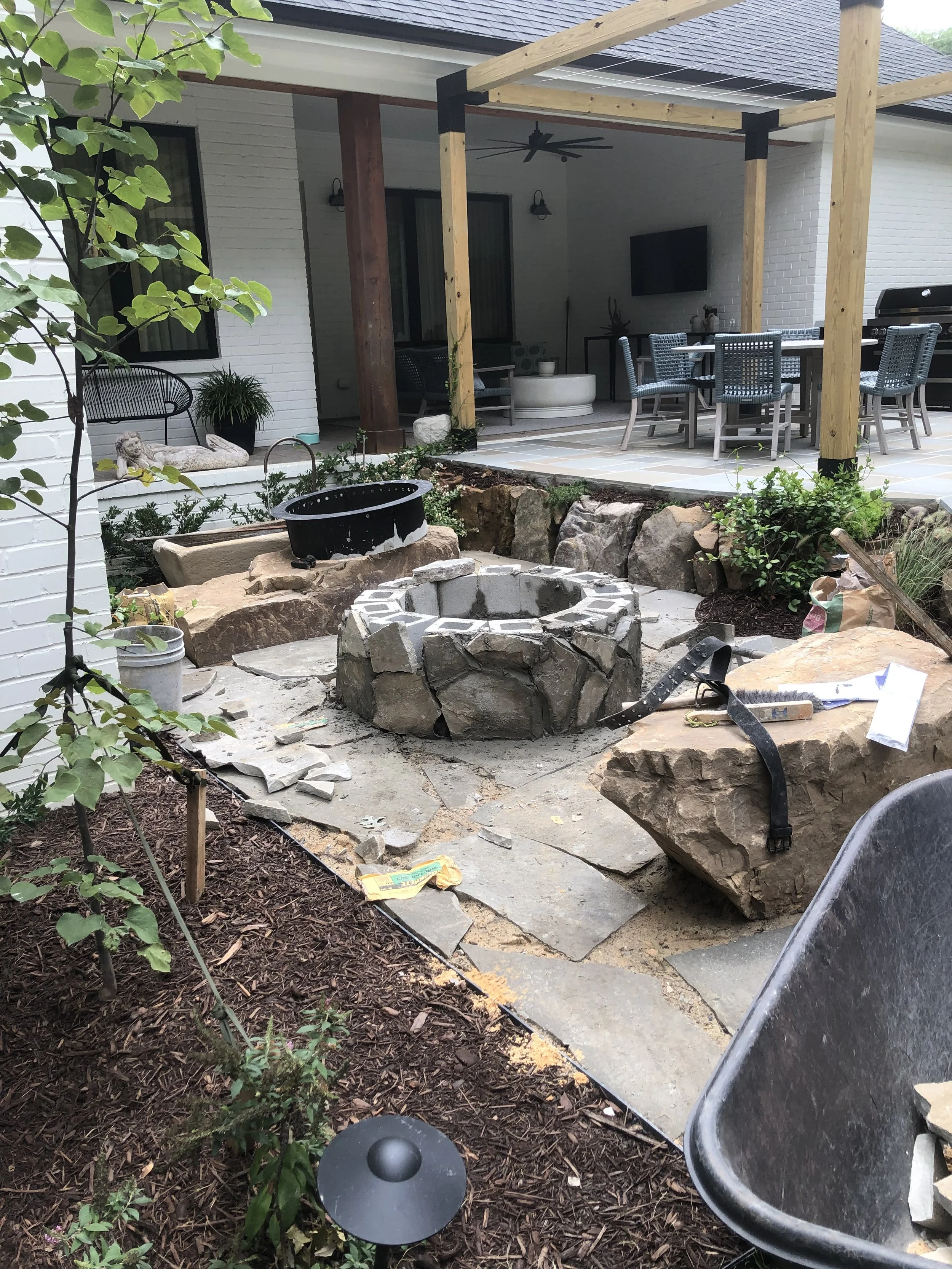A backyard patio area with outdoor furniture, a stone fire pit under construction, plants, and gardening tools. Part of a house with white brick walls and a covered seating area.