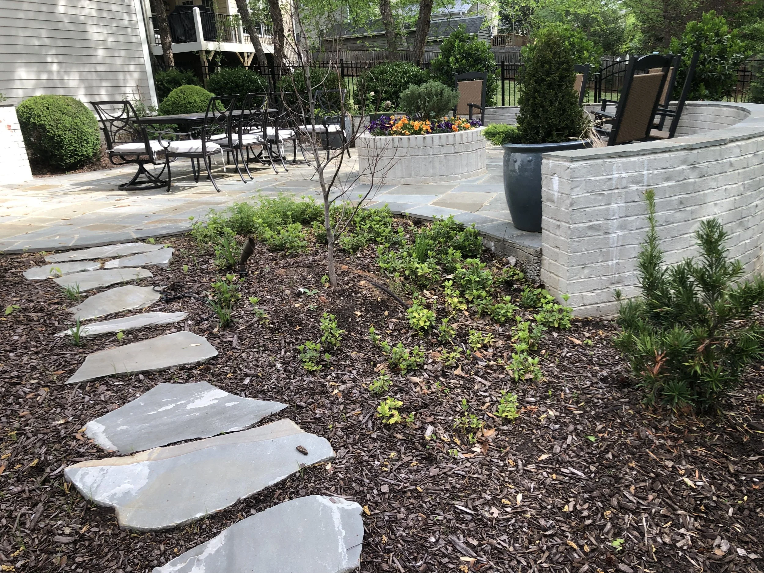 A backyard patio with a stone pathway, a brick retaining wall, garden chairs, a large potted plant, a flower bed with orange and purple flowers, and various shrubs and trees.