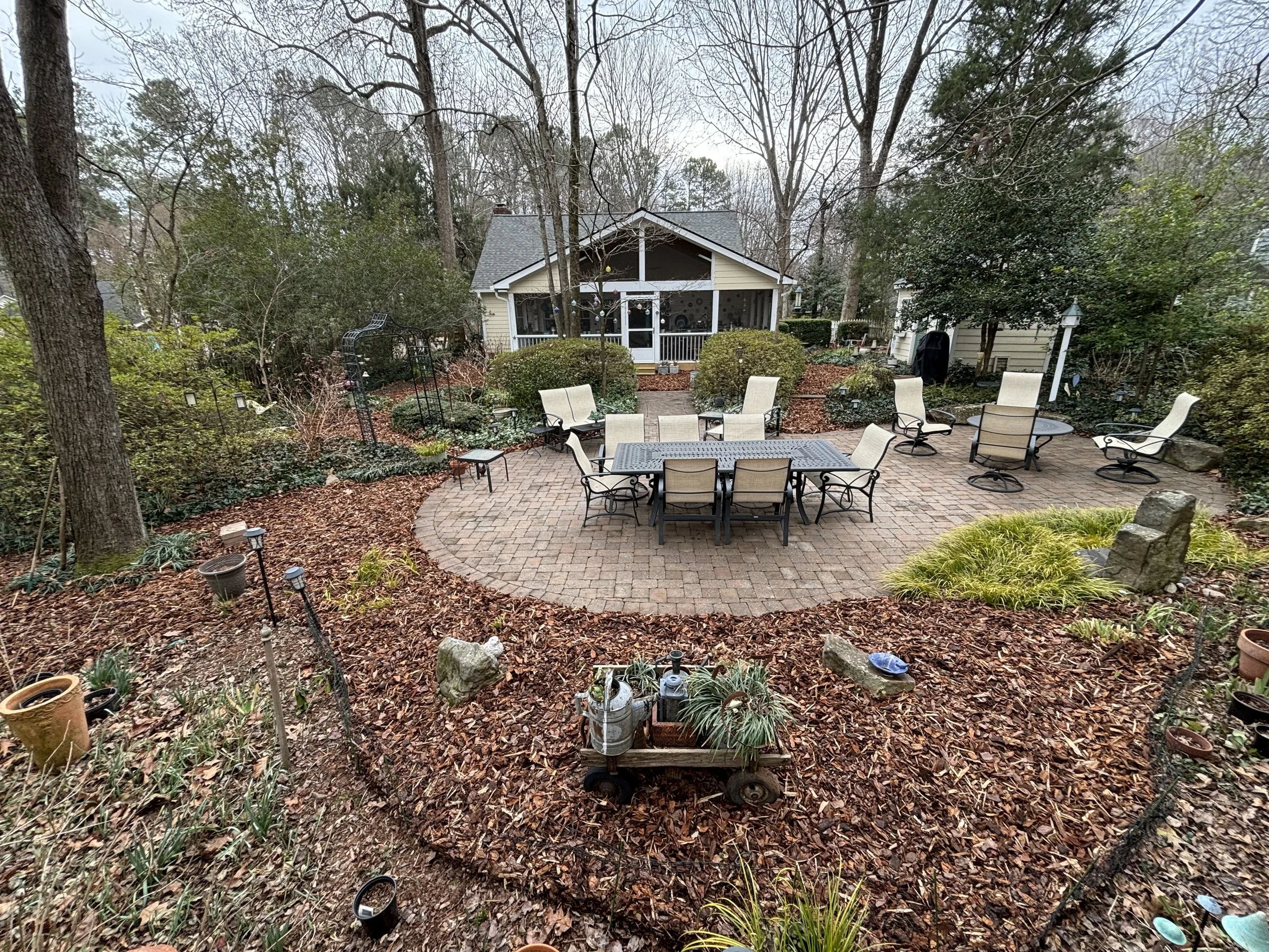 A backyard patio area with a dining table and multiple chairs, surrounded by landscaped bushes, trees, and garden decor, with a house and covered porch in the background.