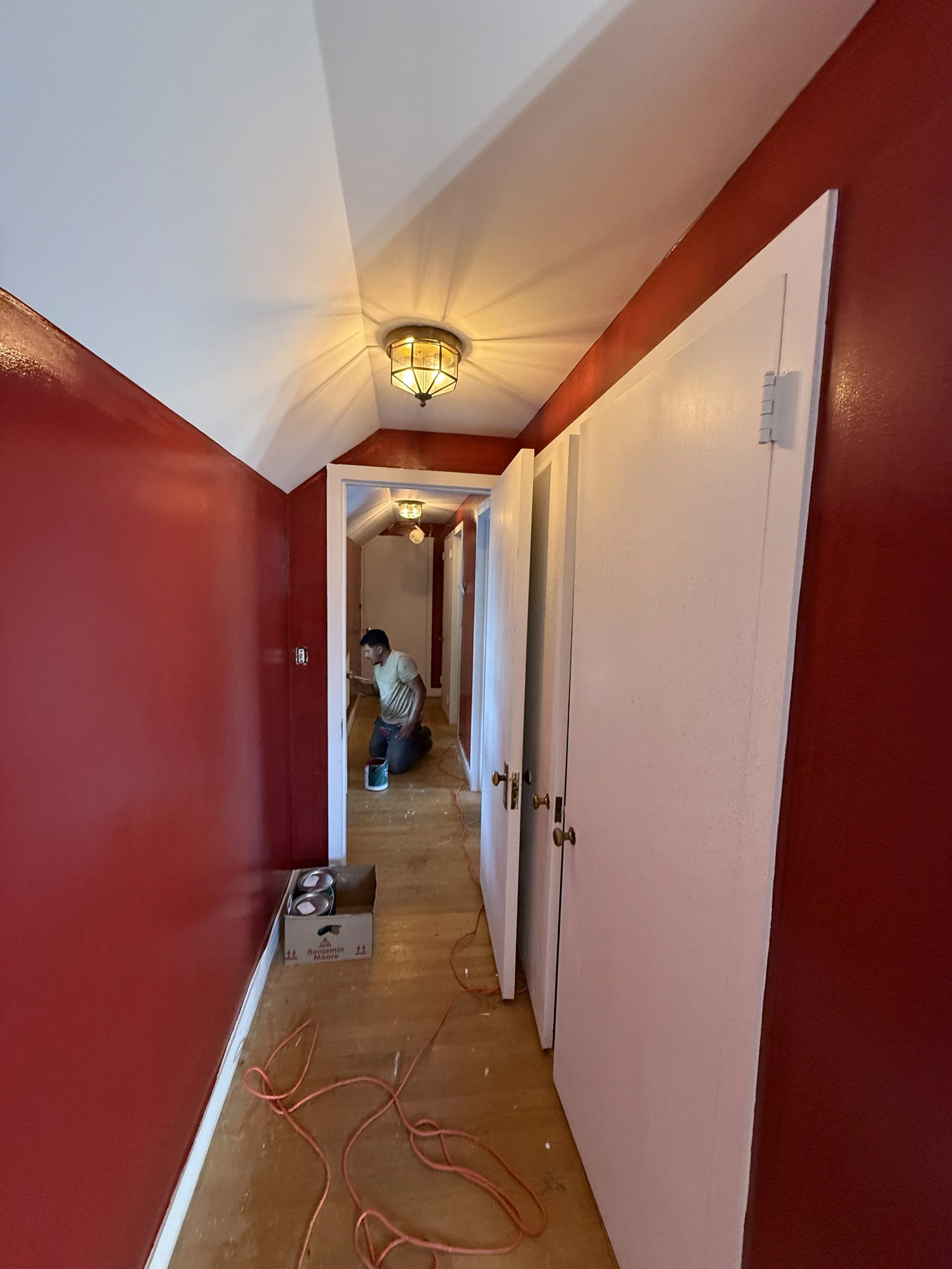 A man kneeling on a wooden floor in a hallway, painting the white ceiling with a paint roller, with red walls, a white door open revealing another room, and an orange extension cord on the floor.