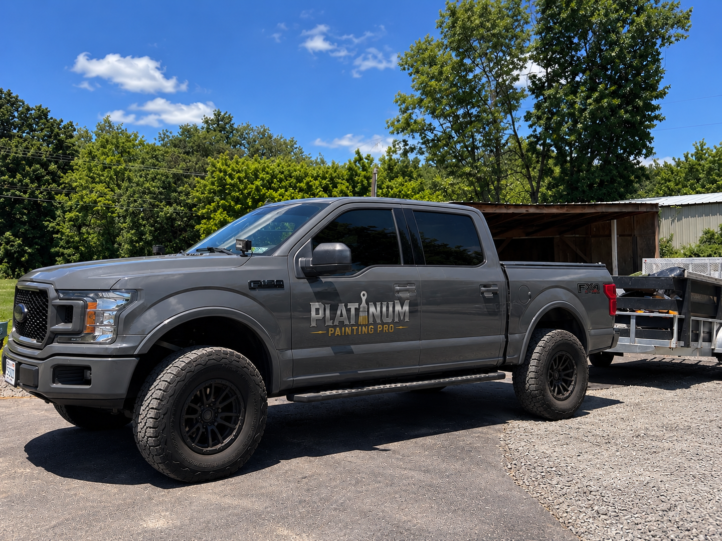 A gray Ford F-150 pickup truck with 'Platinum Painting Pro' logo on the door, parked on a gravel driveway under a blue sky with trees and a wooden shed in the background.