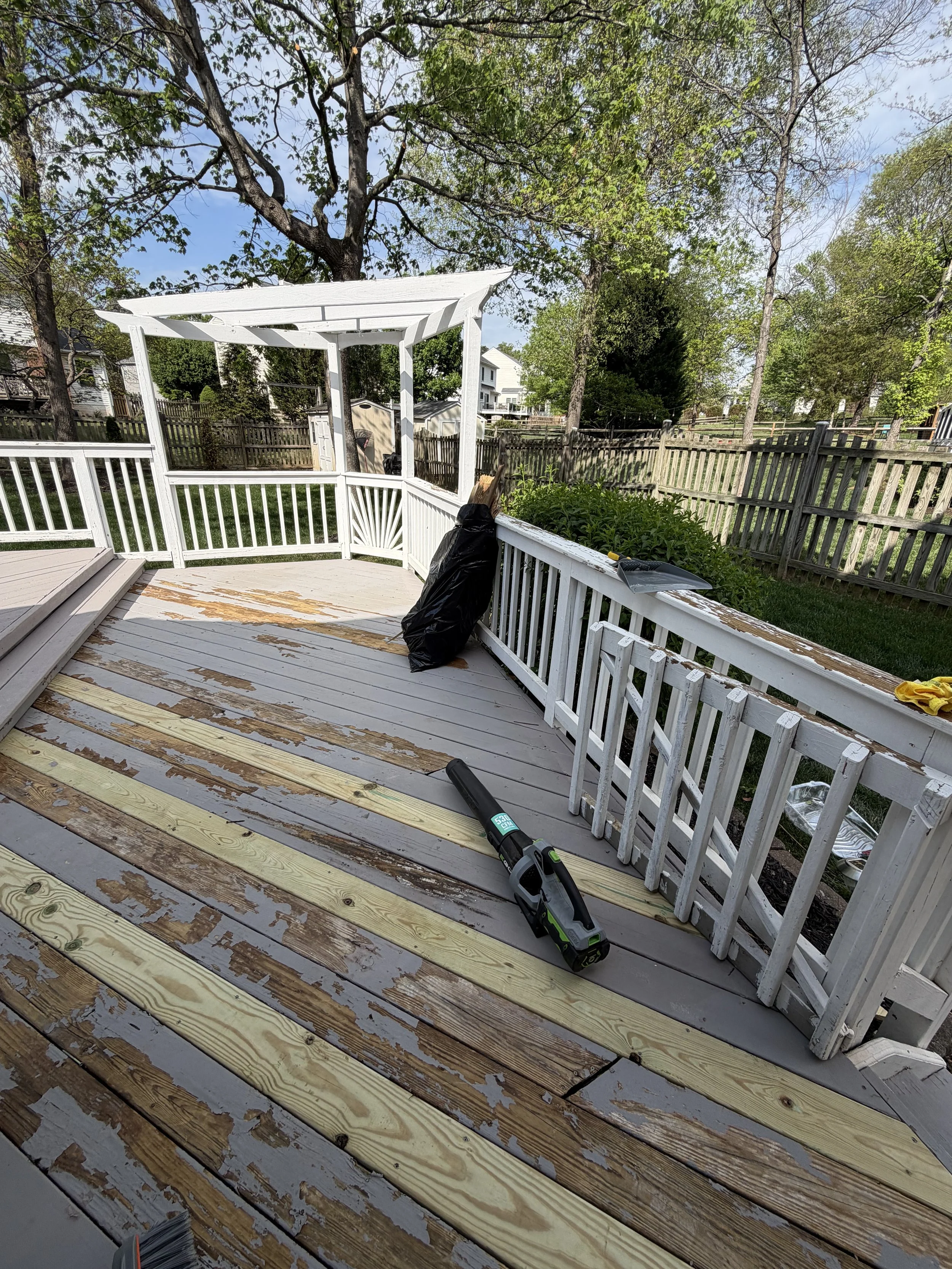 A backyard deck under construction with partially installed wooden floorboards and a white railing. Some tools and materials, including a cordless power drill, a black bag, and yellow gloves, are scattered around. Trees and neighboring houses are see
