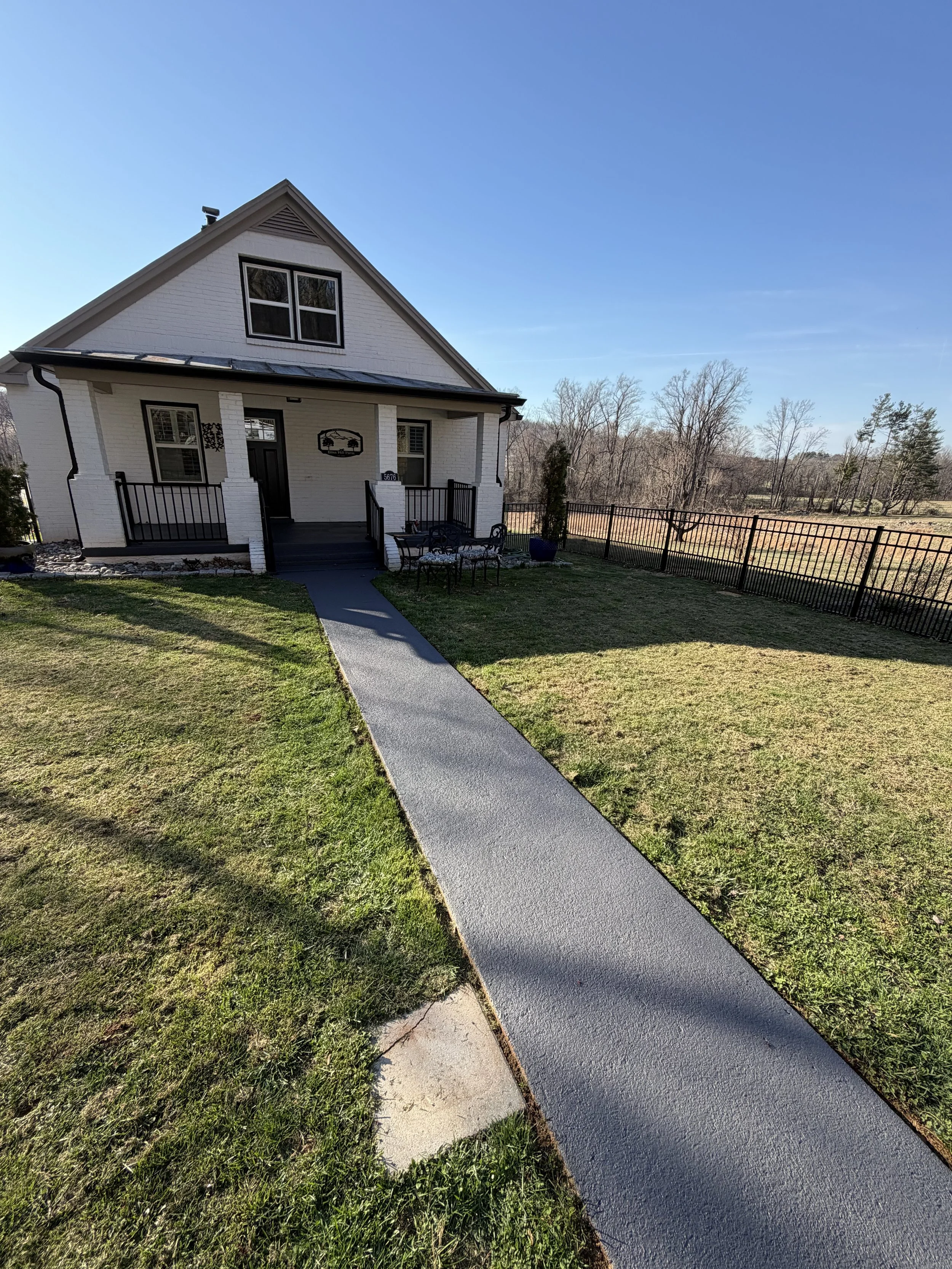 A white two-story house with black window frames and a small front porch with black railing, surrounded by a grassy lawn and a black metal fence, under a clear blue sky.