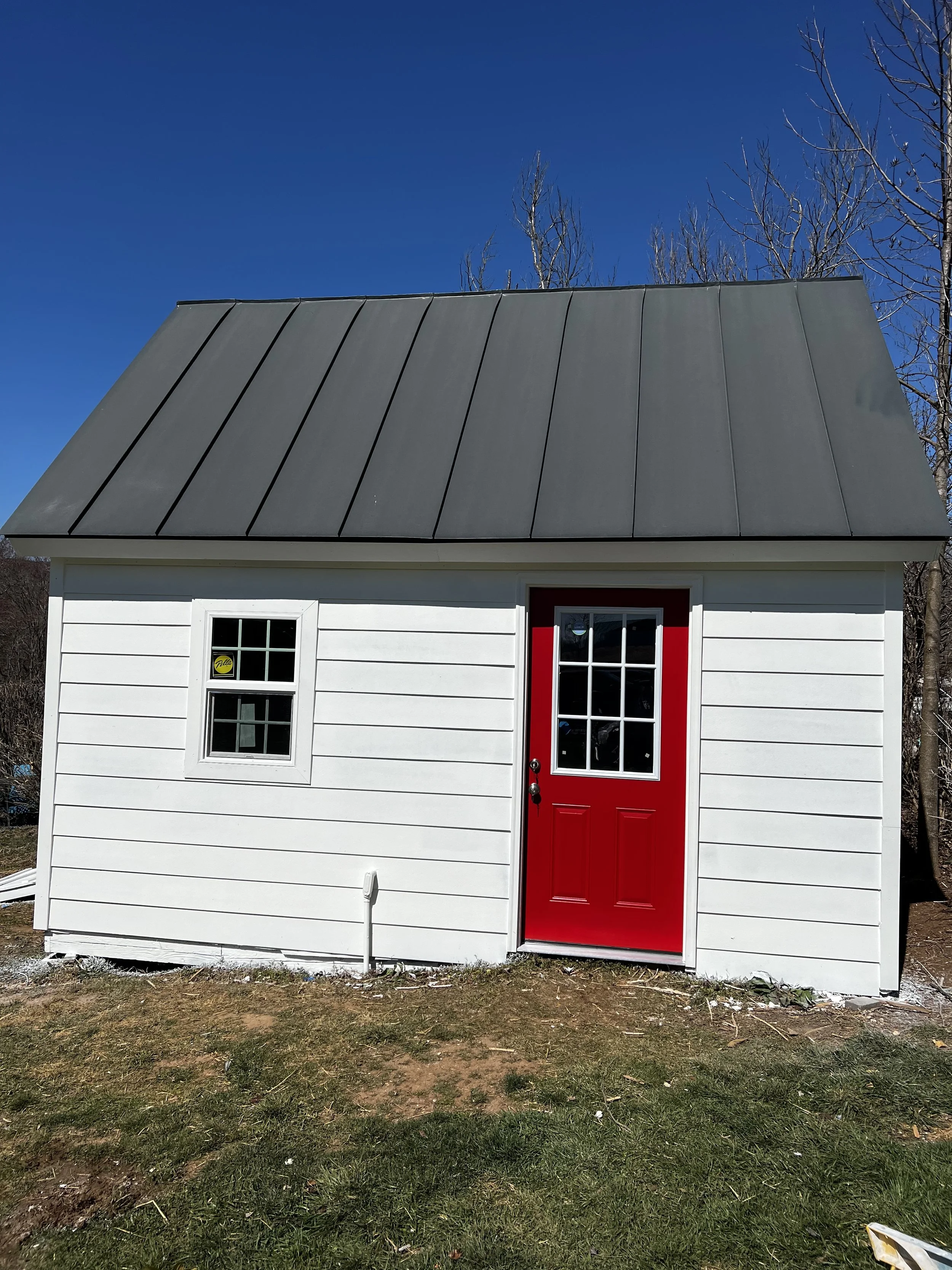 Small white building with a red door and black metal roof, two windows, and a patch of grass in front, under a clear blue sky with a few bare trees in the background.