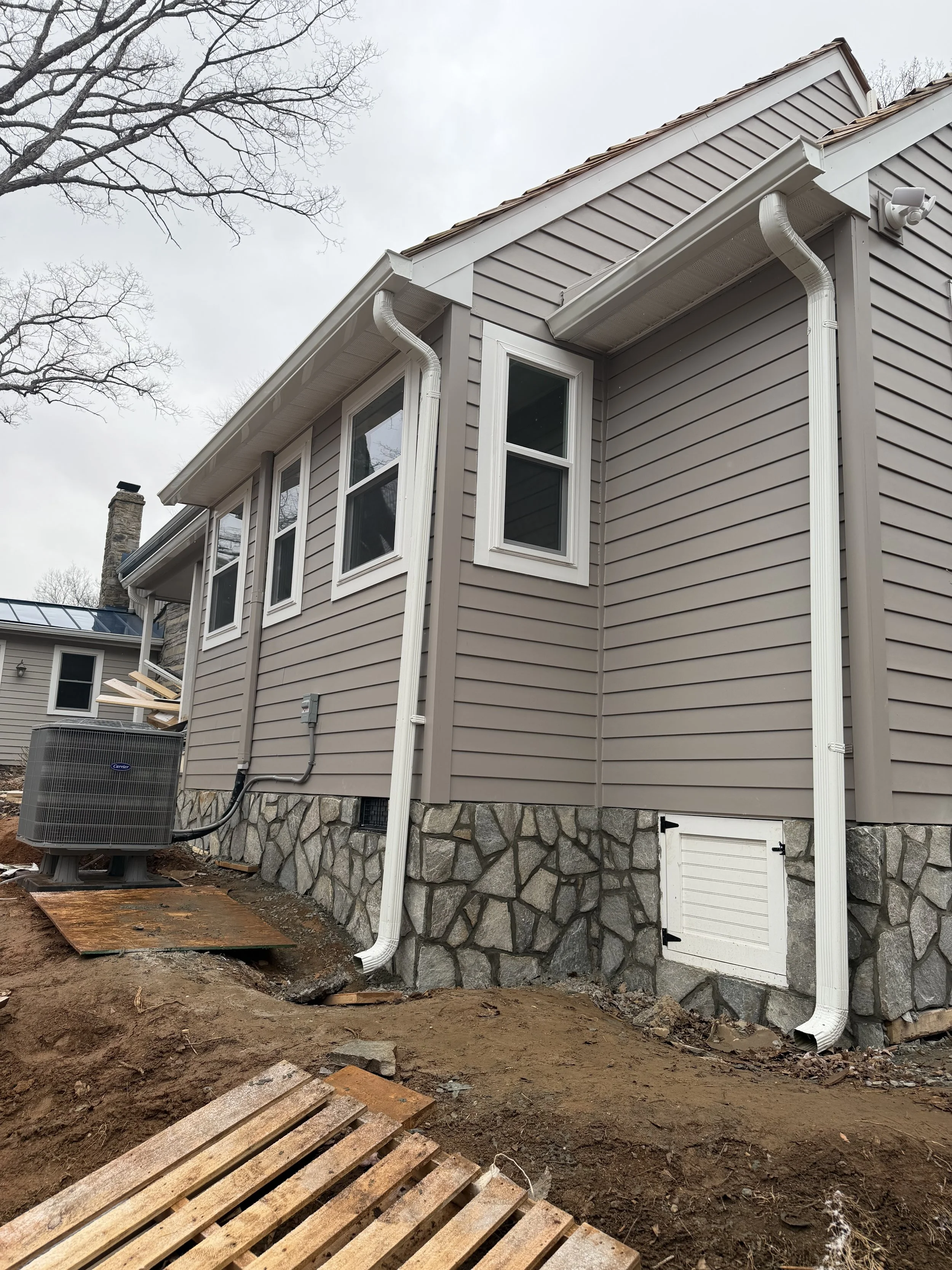 Back of a house with beige siding, three white-framed windows, a stone foundation, and white gutters. An air conditioning unit, construction materials, and exposed ground are visible in the yard, indicating ongoing house construction or renovation.