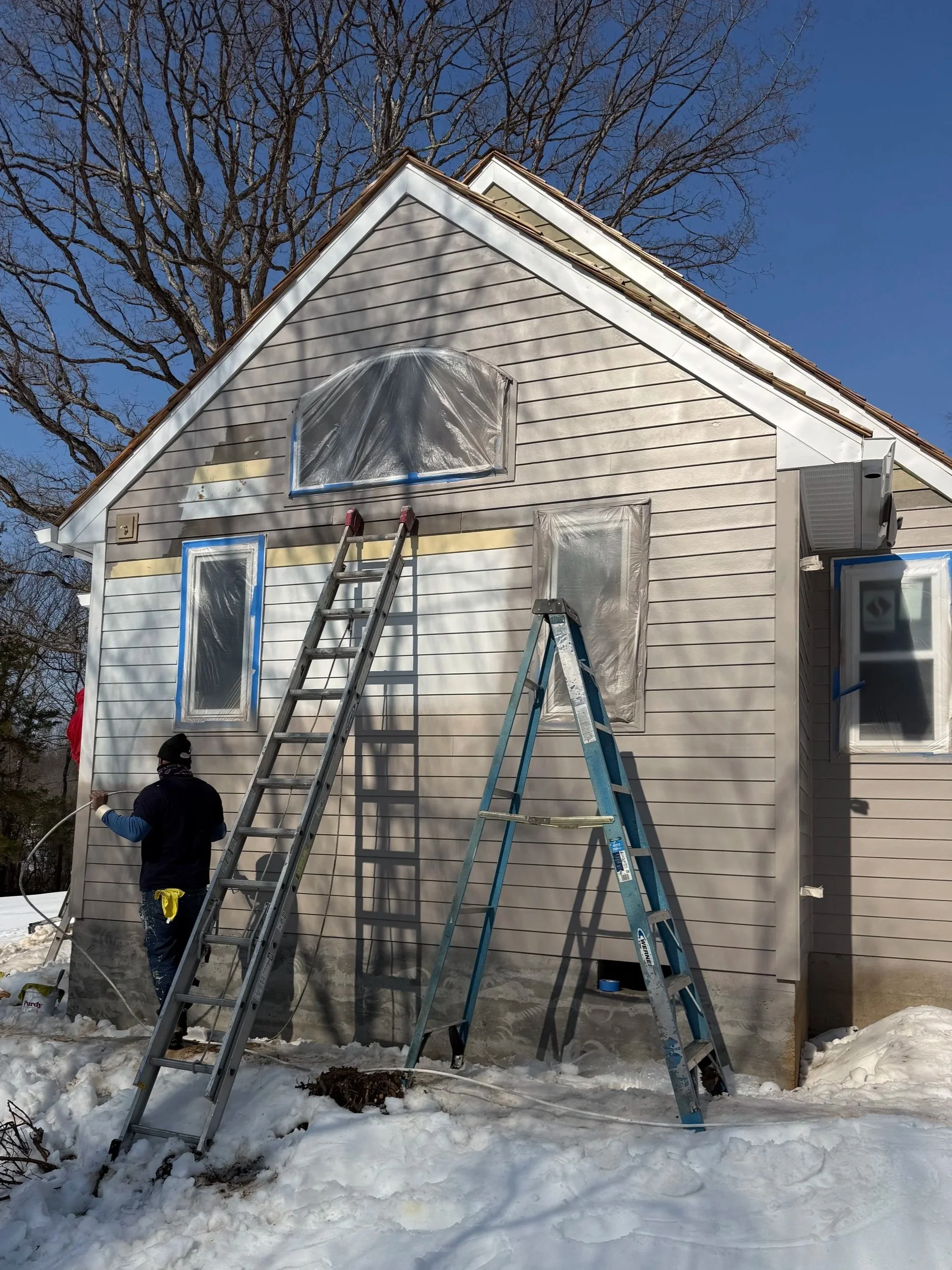 Workers painting the outside of a house during winter, with snow on the ground and ladders leaning against the house.